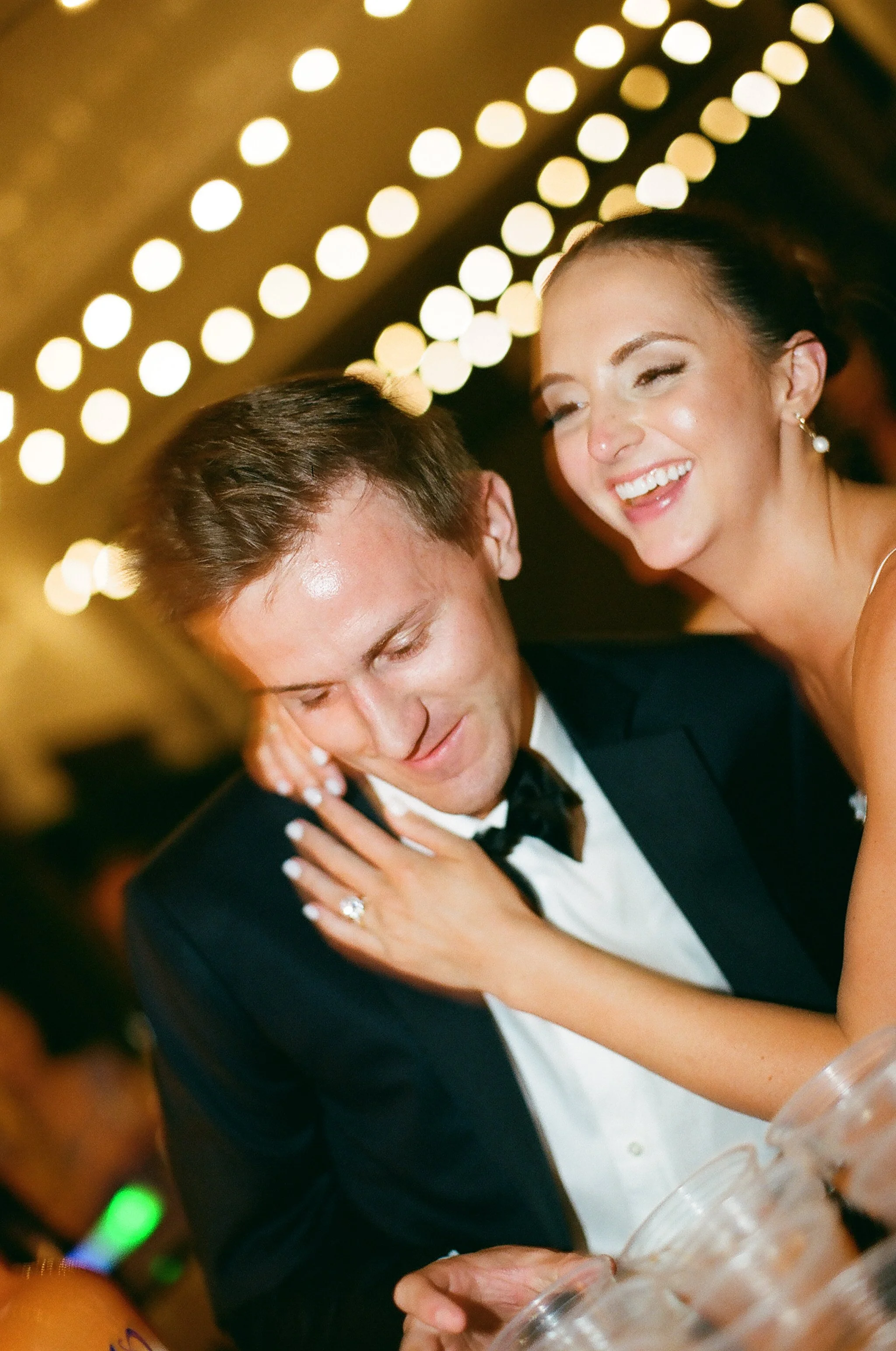 A smiling woman dancing closely with a man in a tuxedo at a celebration or wedding, with blurred warm lights background.