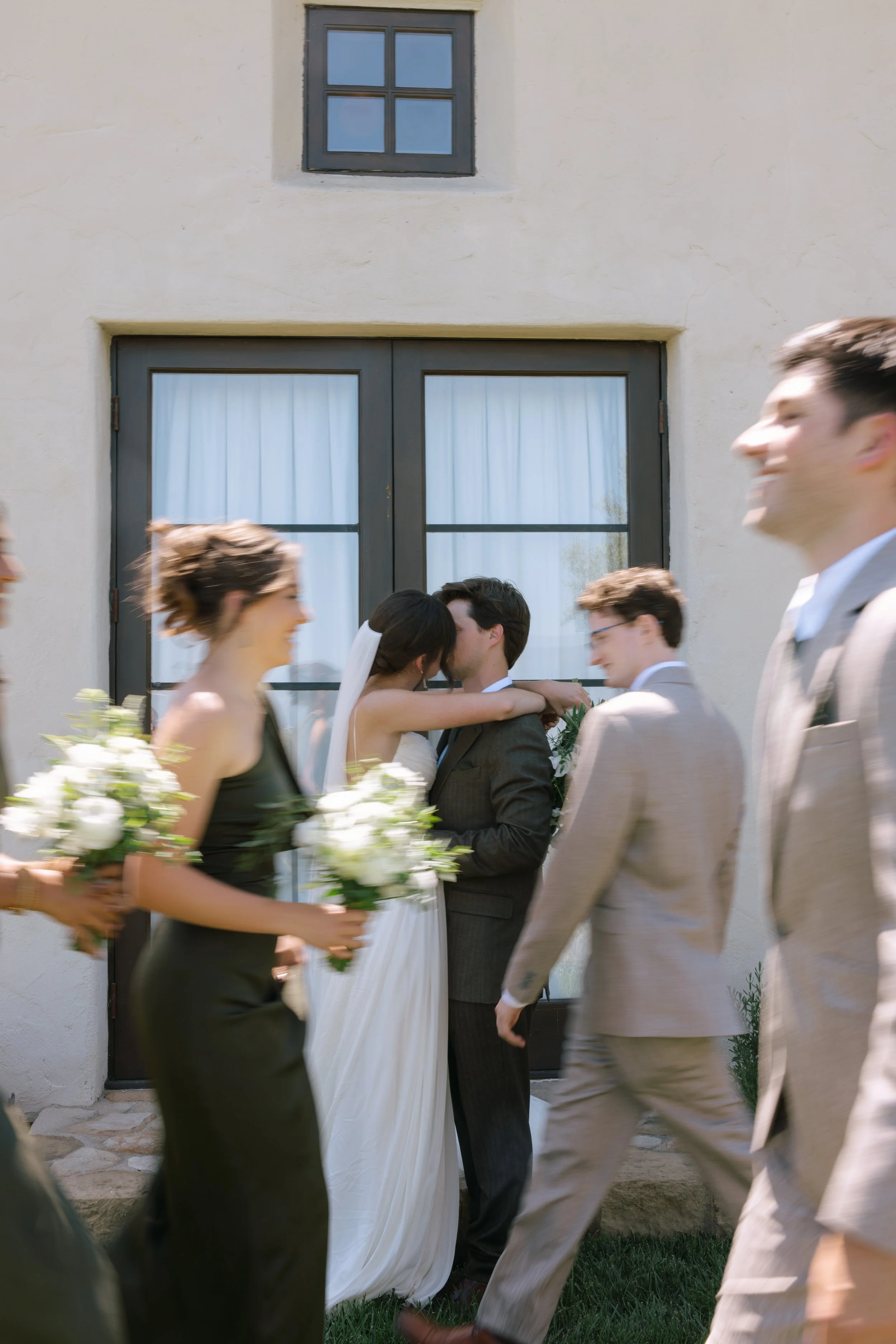 A wedding scene with a bride and groom embracing in front of a beige wall with a small window, surrounded by wedding guests walking in the foreground.