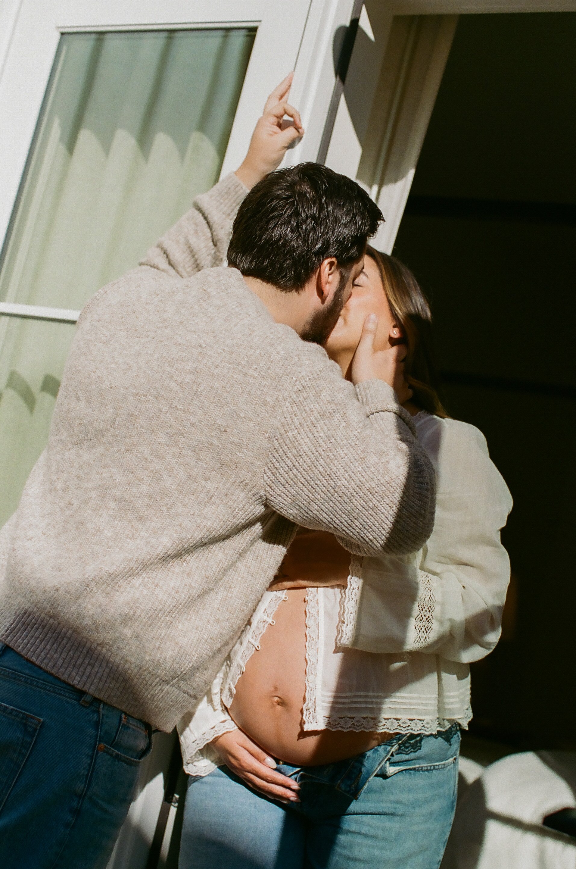 A couple sharing a kiss in a close embrace, with the woman pulling up her shirt to reveal her pregnant belly. They are standing near a door with a glass window.