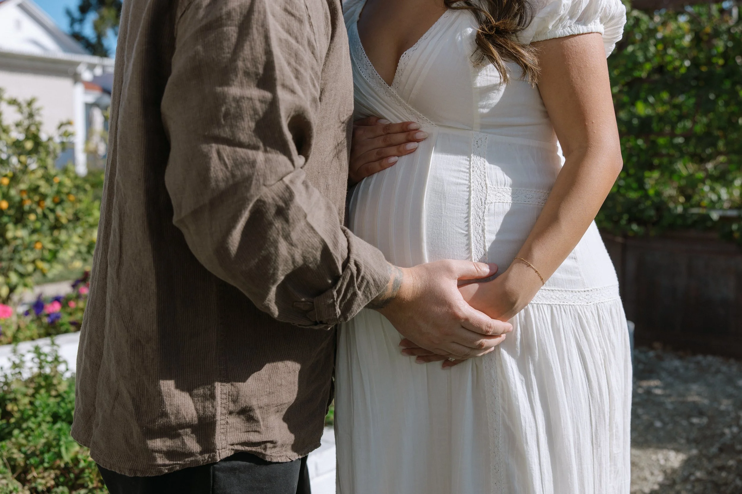 A pregnant woman in a white dress is being touched by a person in a brown shirt, both outdoors in front of greenery.