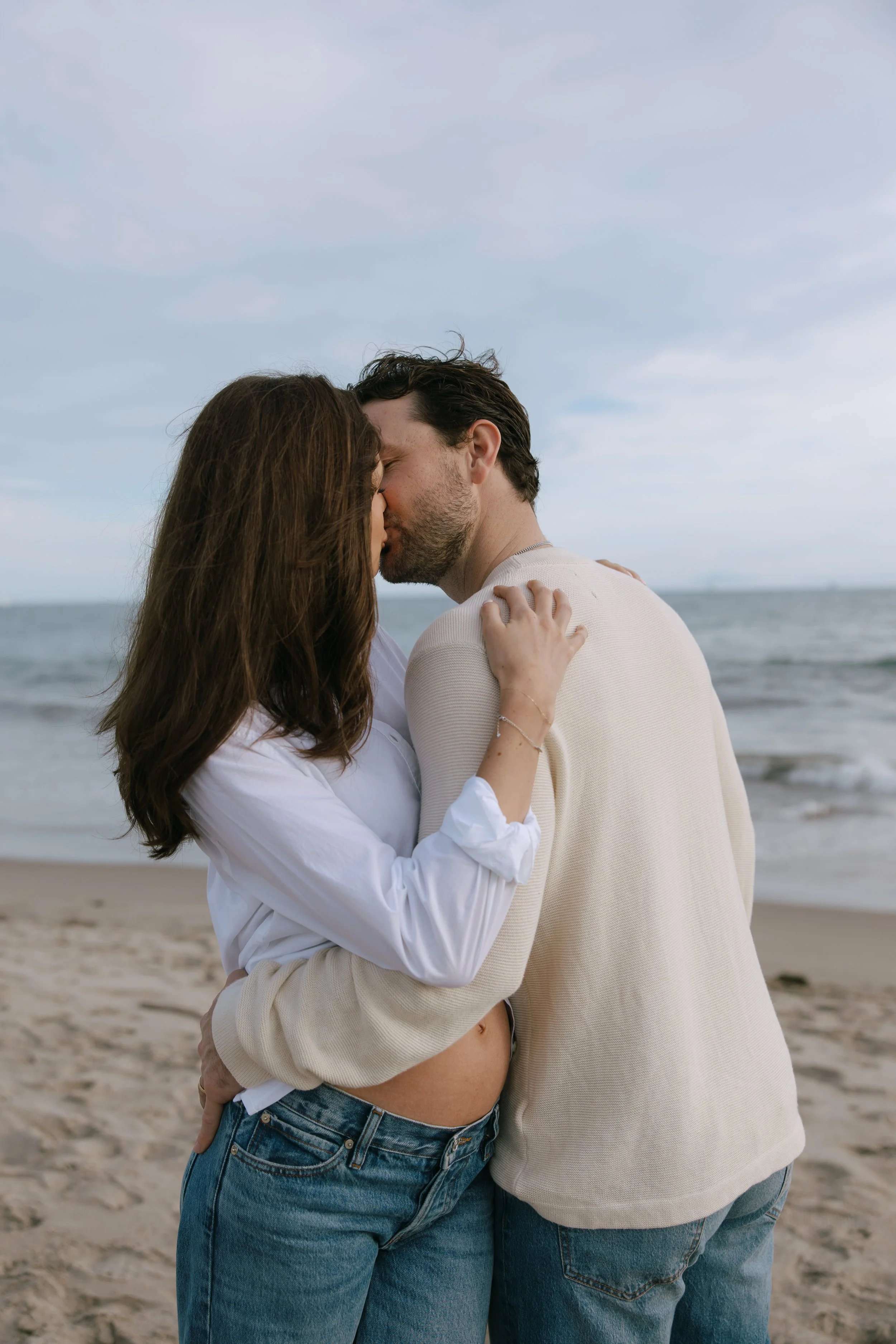 A couple is embracing and kissing on the beach with the ocean in the background, both wearing casual clothing.