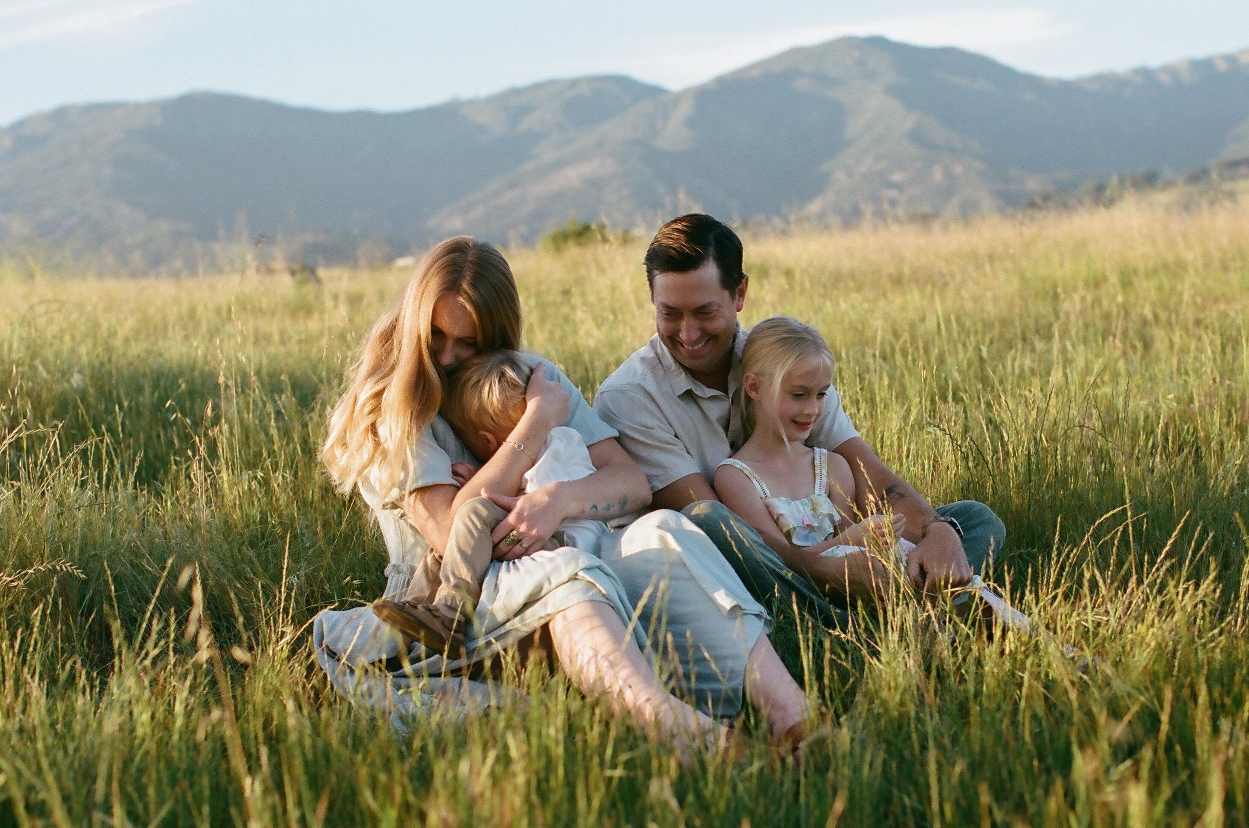 A family of four sitting in a grassy field with mountains in the background, smiling and enjoying time together.