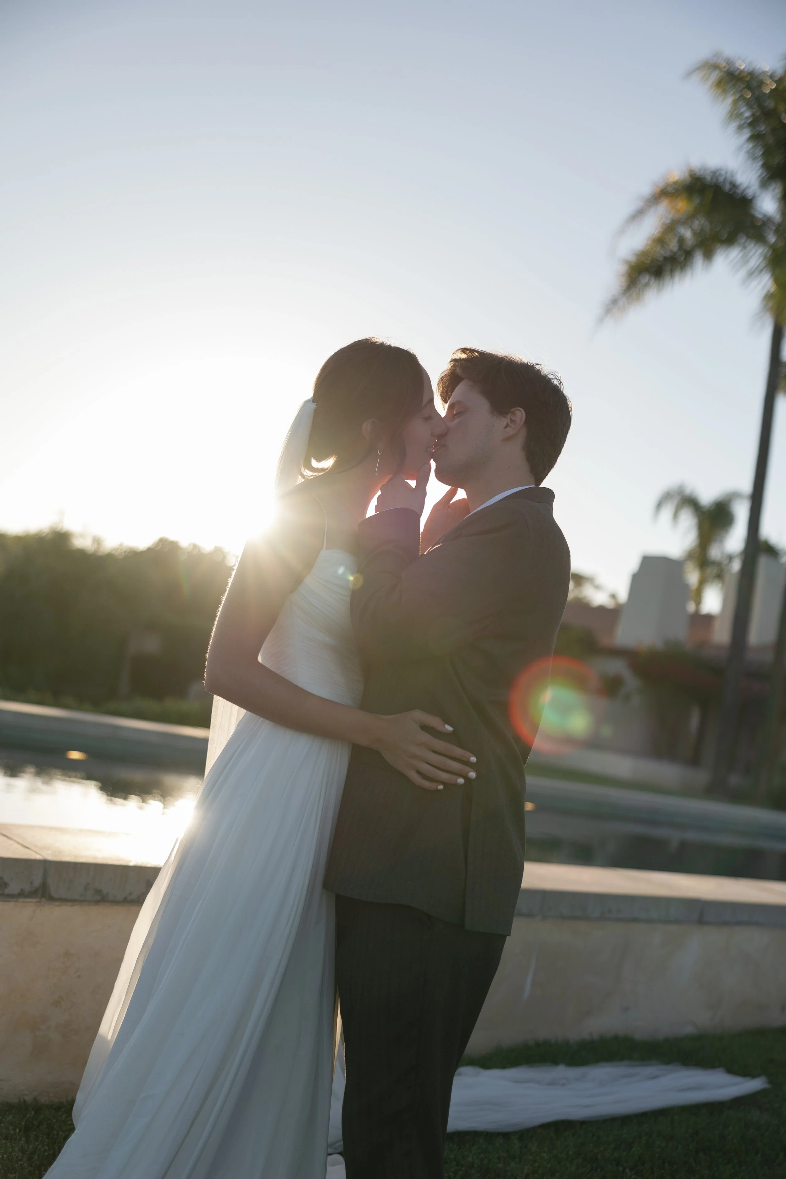 Couple in wedding attire sharing a kiss outdoors at sunset, palm trees in background.