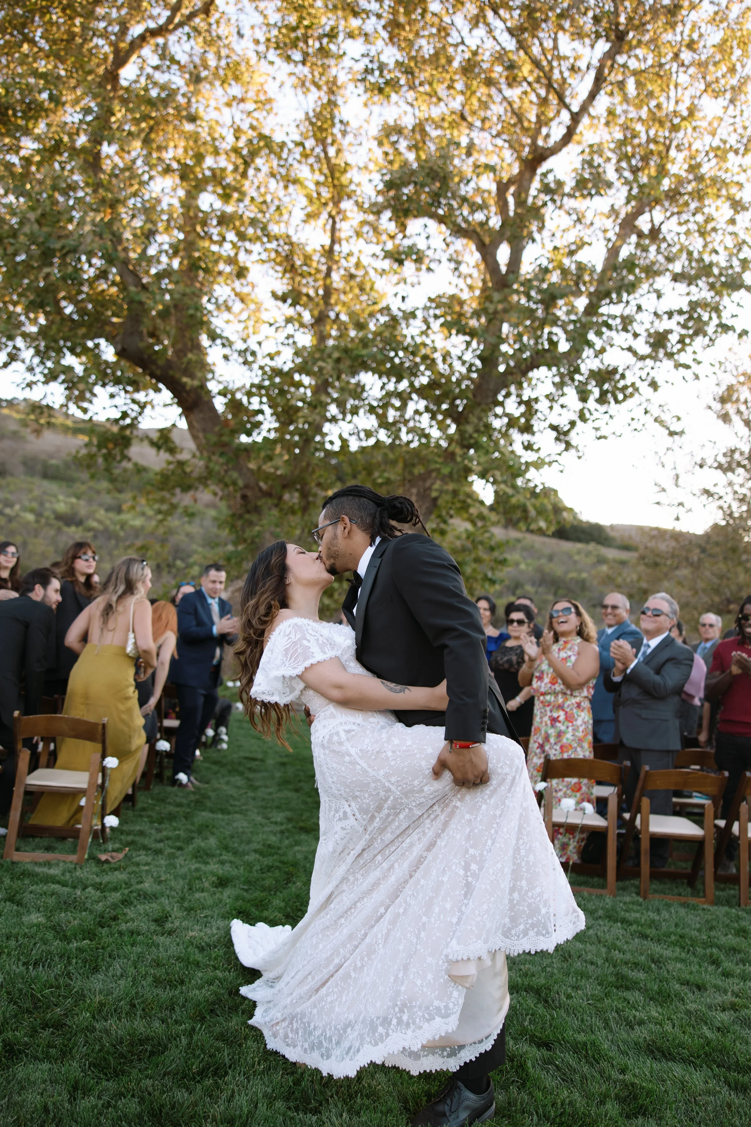 A newlywed couple shares a kiss during their outdoor wedding ceremony on a grassy field, with guests watching in the background under a large tree with autumn-colored leaves.