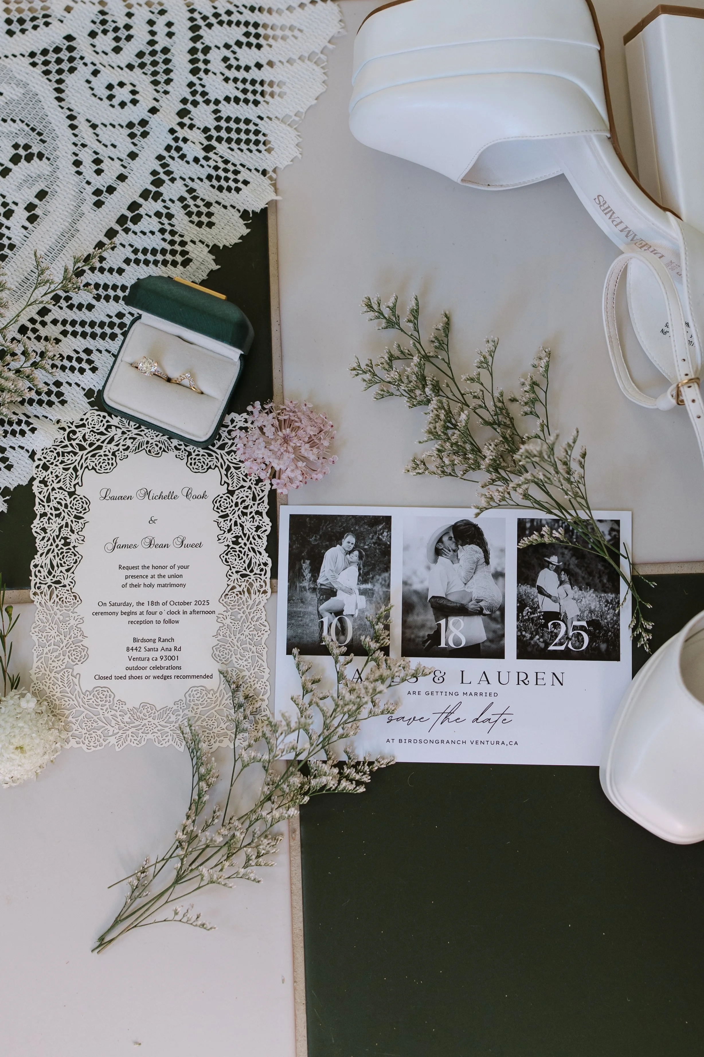 Wedding invitation suite, including an ornate lace wedding invitation, a black and white photo save-the-date card with a couple, a green jewelry box with rings, white flowers, and decorative greenery on a white and black surface.