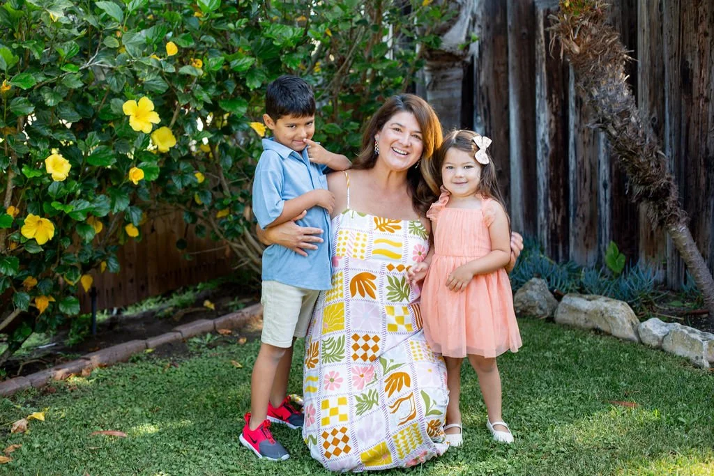 A woman with two children standing outdoors near a wooden fence and yellow hibiscus flowers. The woman is smiling, wearing a colorful dress. The girl is in a peach dress with a bow in her hair, and the boy is in a blue shirt with beige shorts, and red and black shoes.