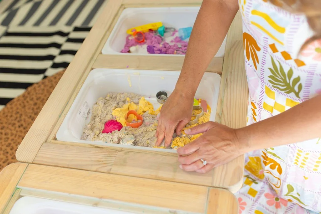Person playing with colorful kinetic sand and toys in a sandbox on a wooden table.