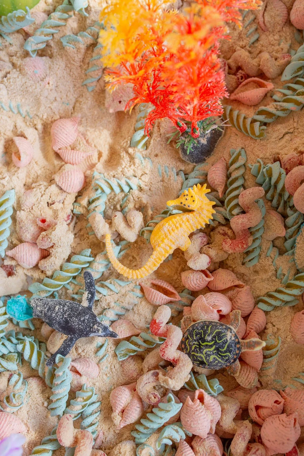 Colorful plastic toy sea creatures including a yellow seahorse, a black shark, an orange coral, a green turtle, and a yellow seahorse, scattered among pink seashells, spiral pasta, and sand.