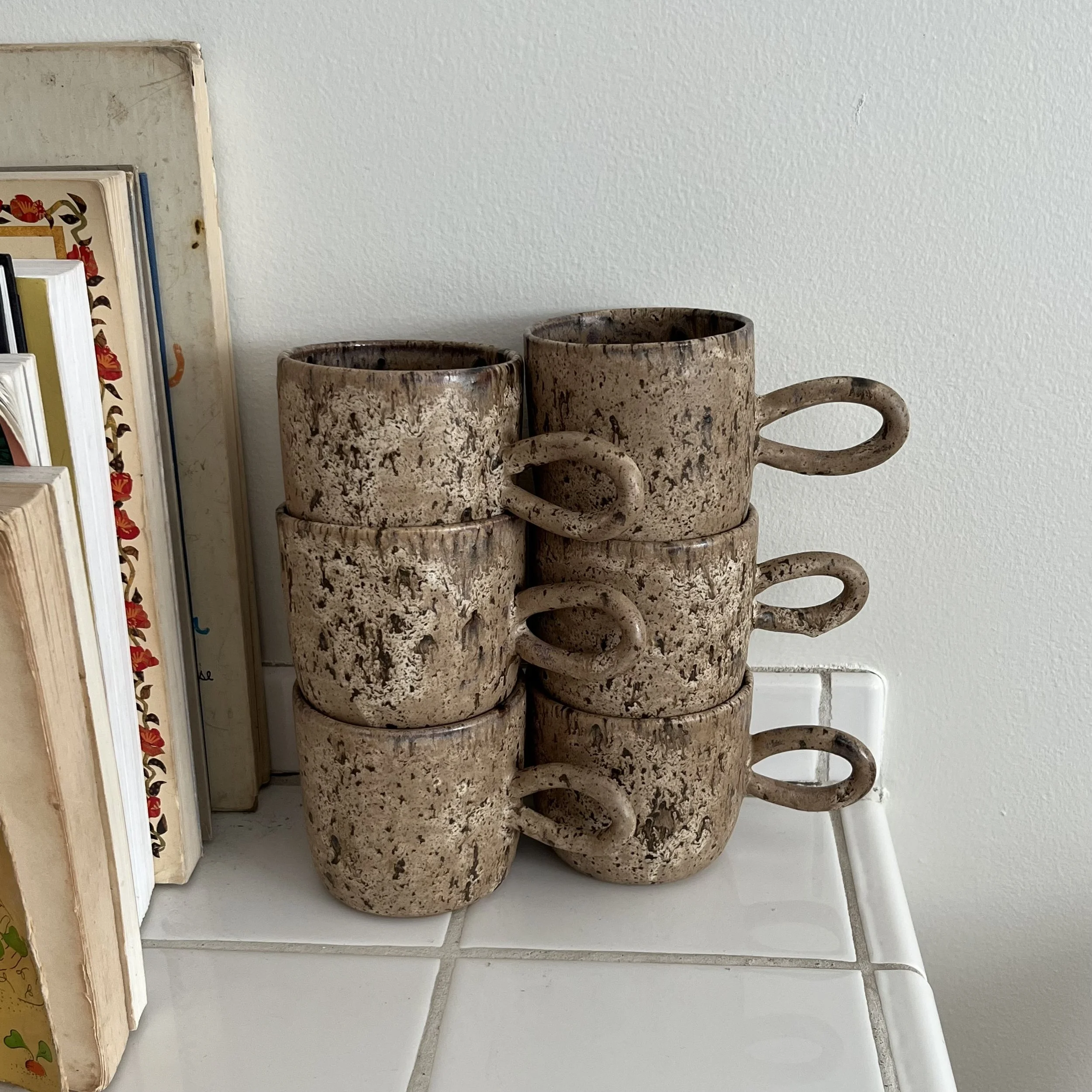 Stacked brown speckled ceramic mugs on a white surface in front of a white wall.