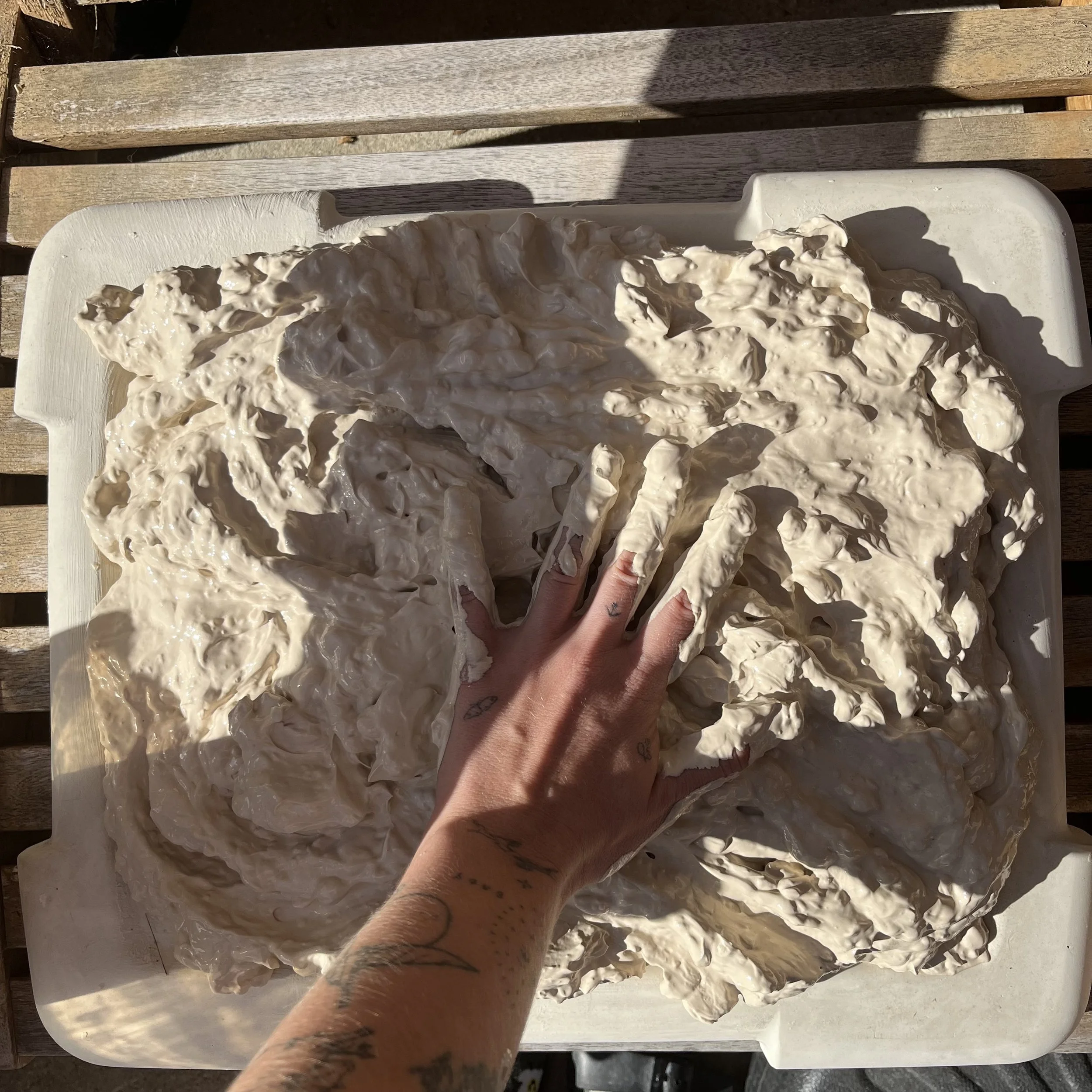 A person's hand and forearm, with tattoos, pressing on a large, uneven white substance on a rectangular white tray, set on a wooden surface outside.