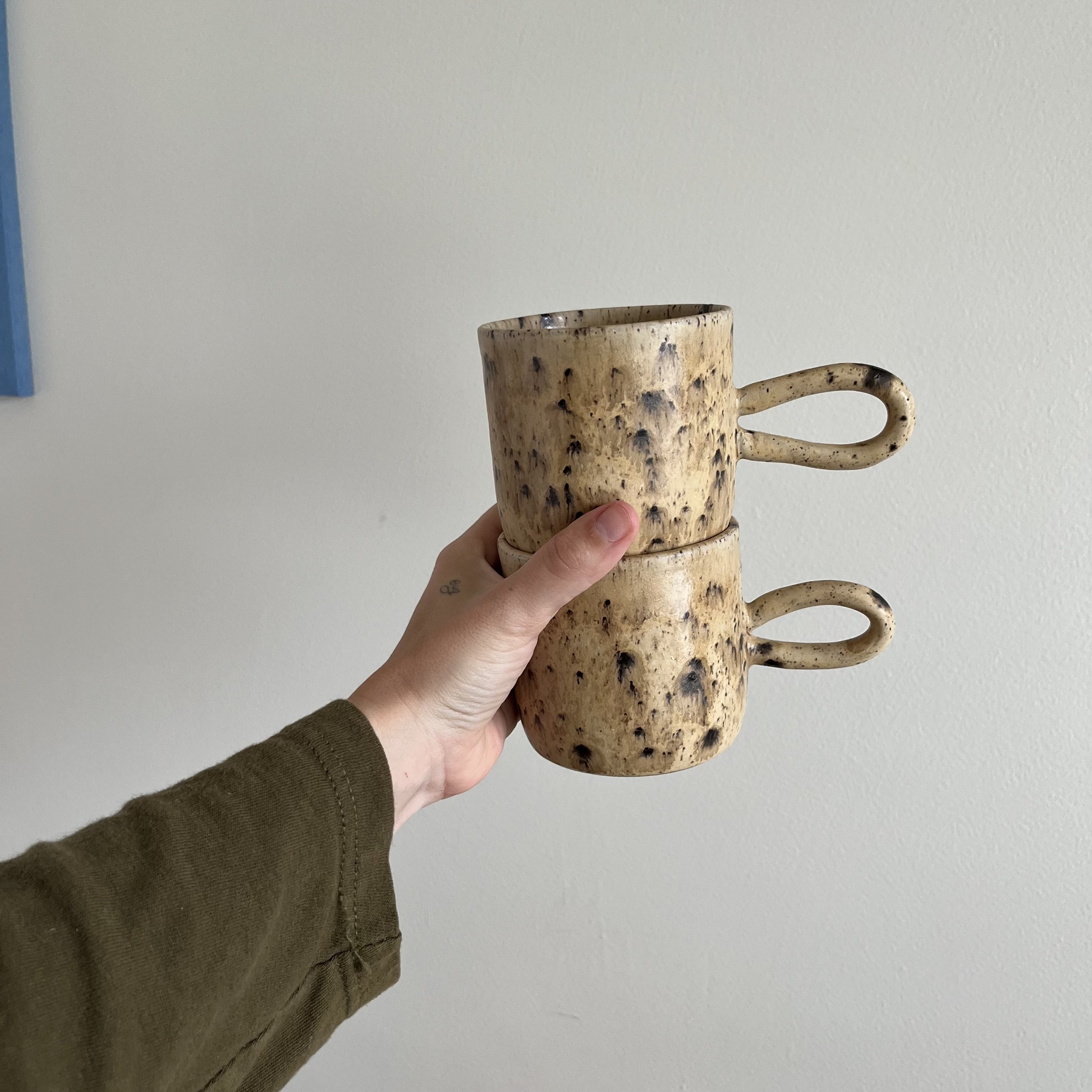 A hand holding a stack of beige speckled ceramic cups in front of a plain white wall.