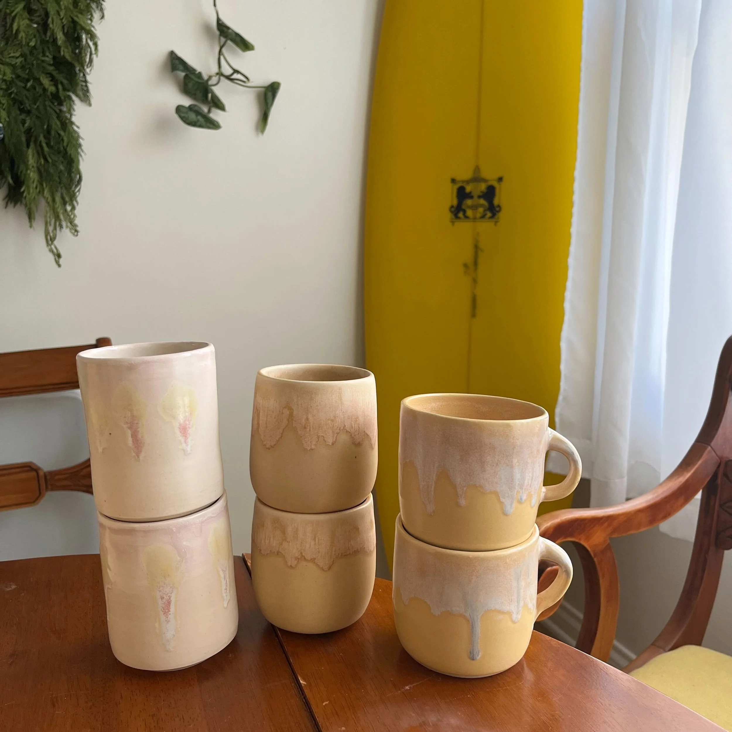 A set of cream-colored ceramic mugs with pink and brown marbling, arranged on a wooden table, with green plants and a yellow curtain in the background.