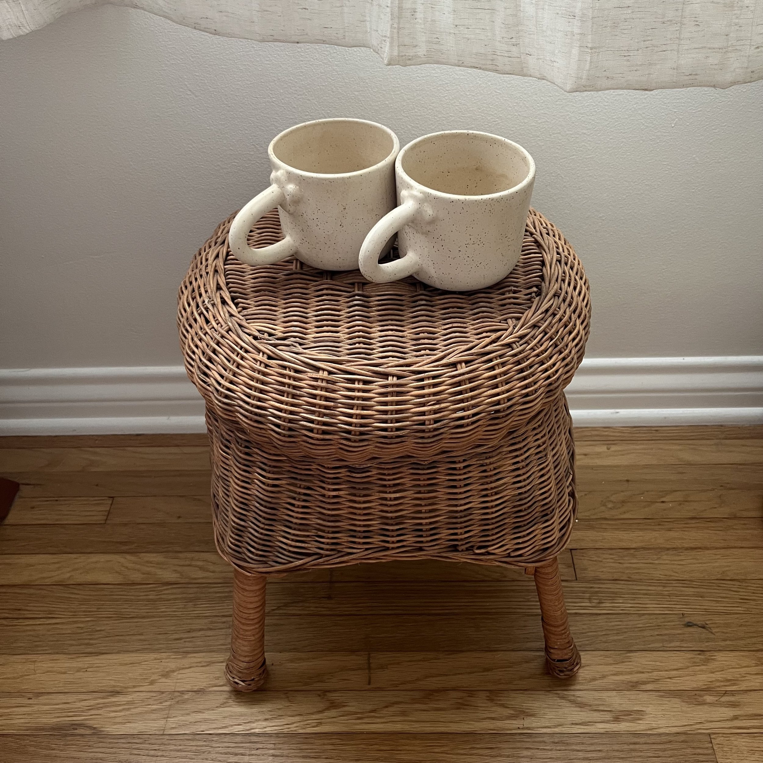 Two cream-colored ceramic mugs with speckled glaze on a round rattan table with four legs, placed against a plain gray wall and a wooden floor.