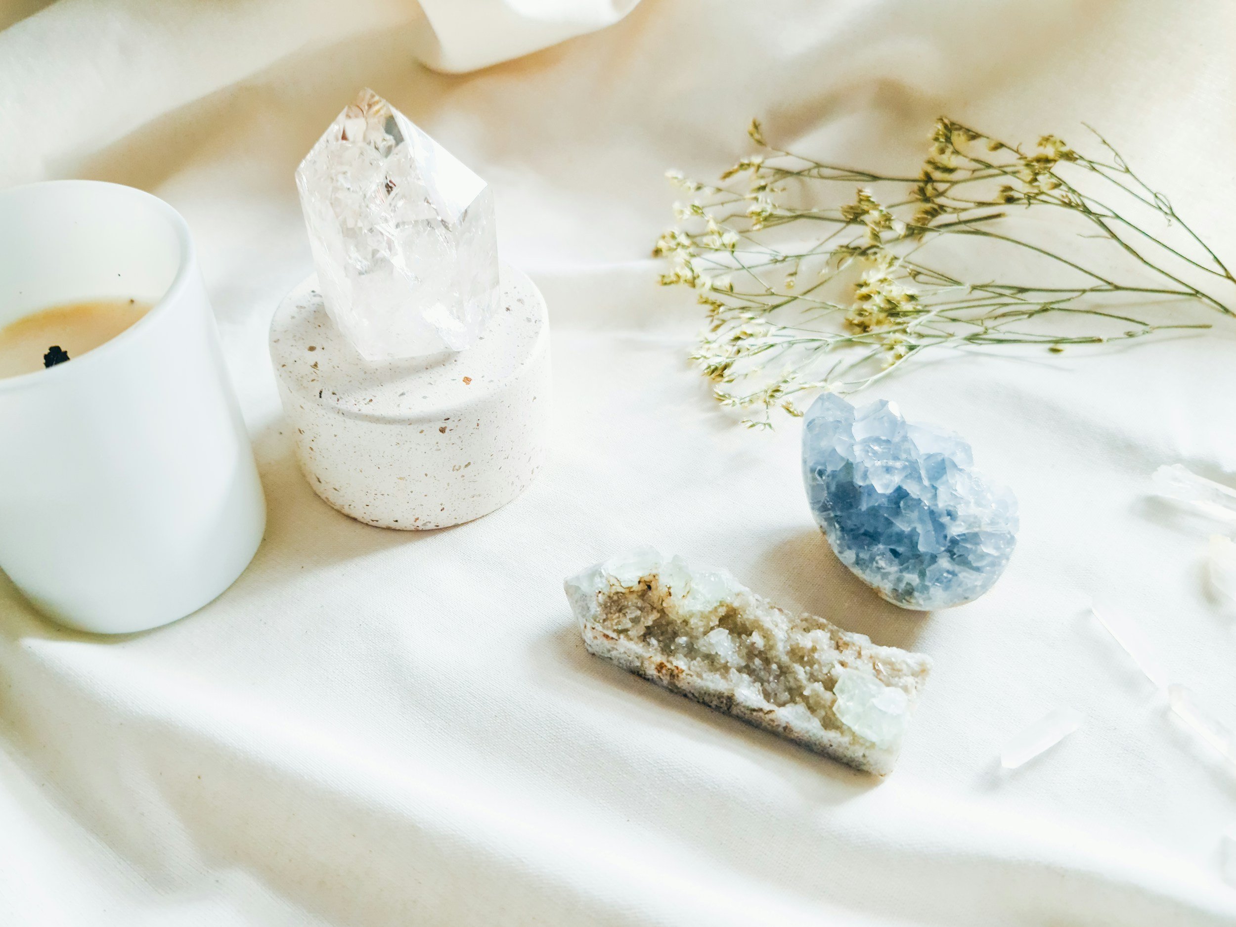 White table with a white mug, a clear crystal on a speckled white stand, a cluster of greenish-white dried flowers, a blue geode, and a white textured mineral specimen.