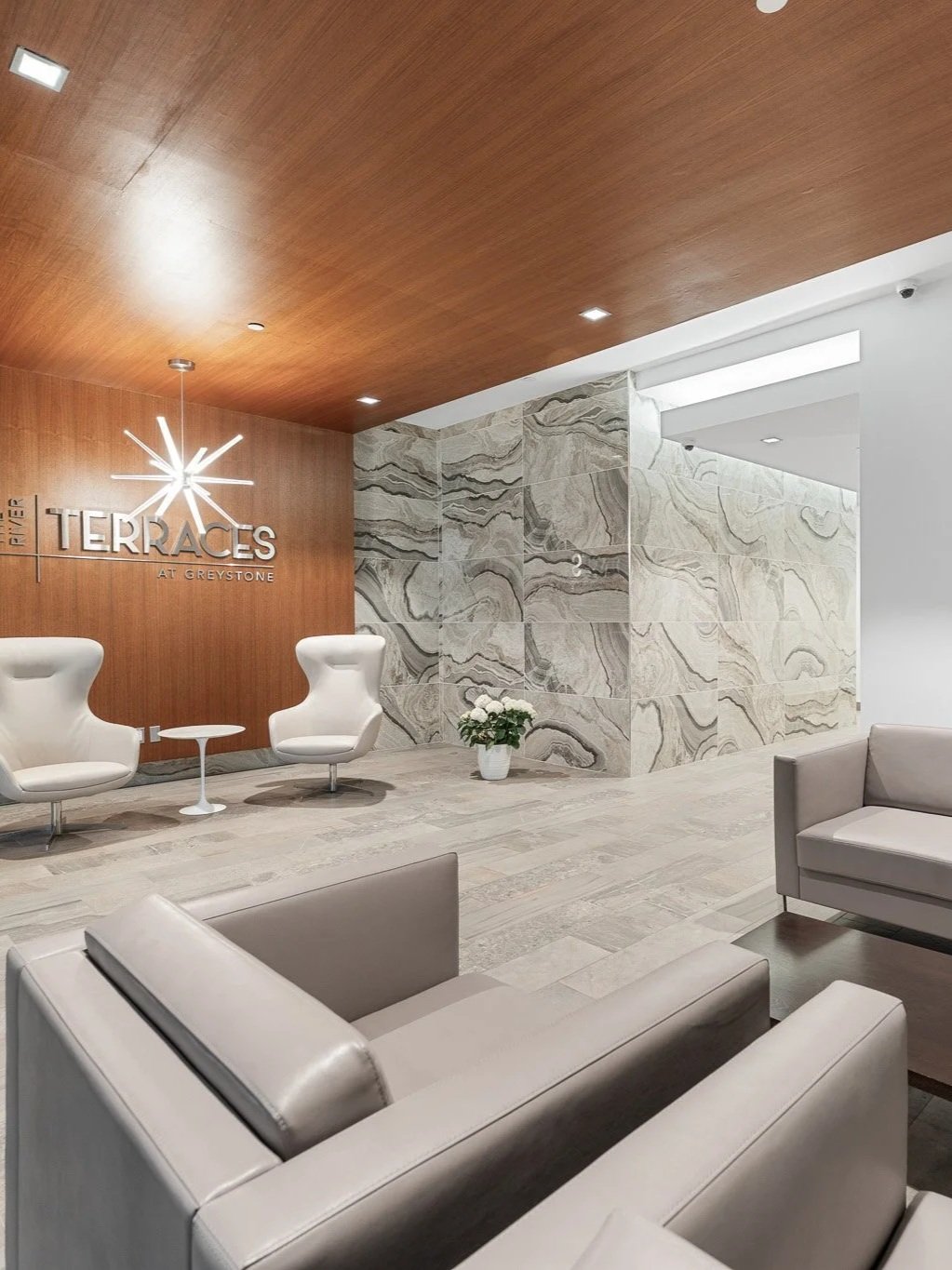 Modern lobby with white armchairs, gray sofas, a small round table, a wooden ceiling, a marble wall, and a sign that reads 'FIVE TERRA CES AT GREYSTONE' illuminated with a starburst light.