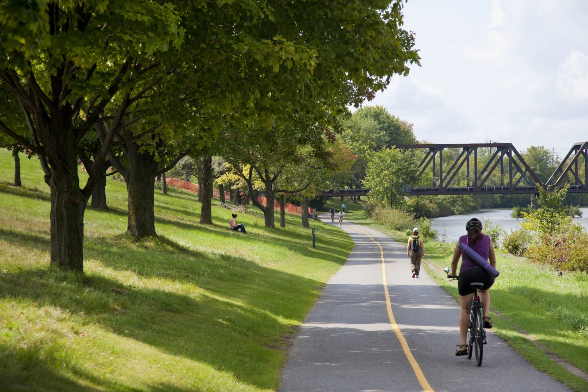 A scenic park pathway with lush green trees on one side and a river on the other, where several people are walking and biking on a sunny day.