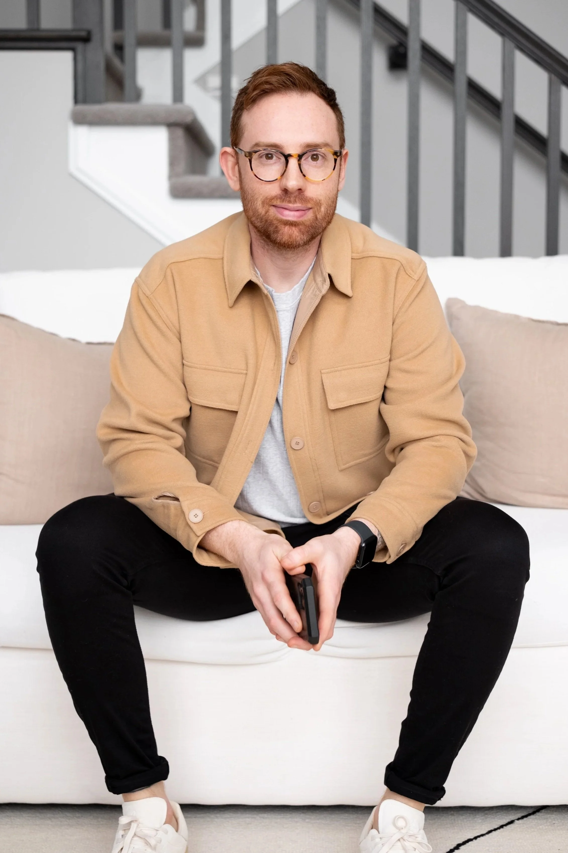 A man with glasses, a beard, and wearing a beige jacket, black pants, and white sneakers sitting on a white sofa holding a smartphone in a living room with a staircase in the background.