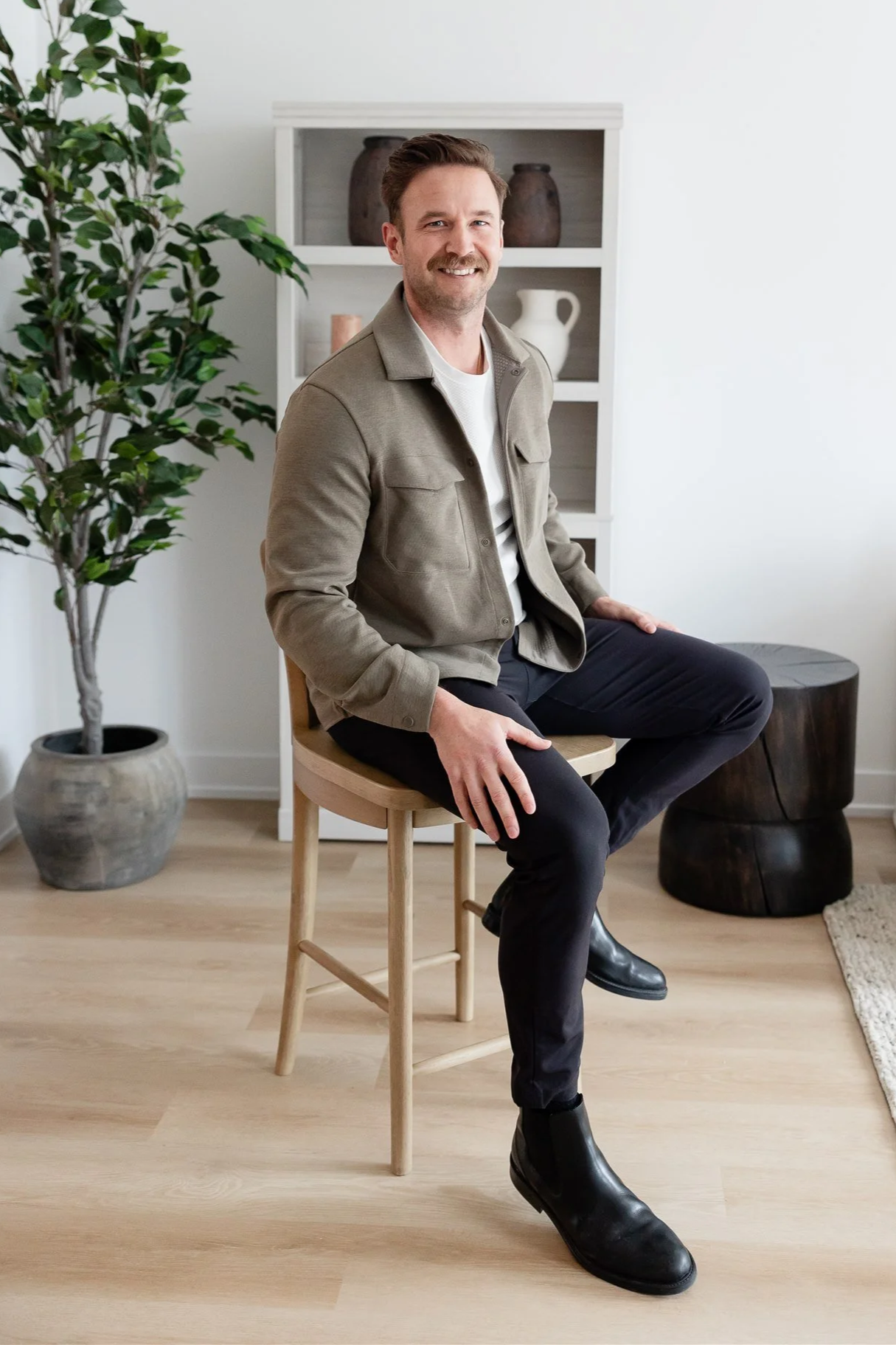 A man sitting on a wooden chair in a modern, minimalistic room with a large potted plant and a white bookshelf with decorative items behind him.