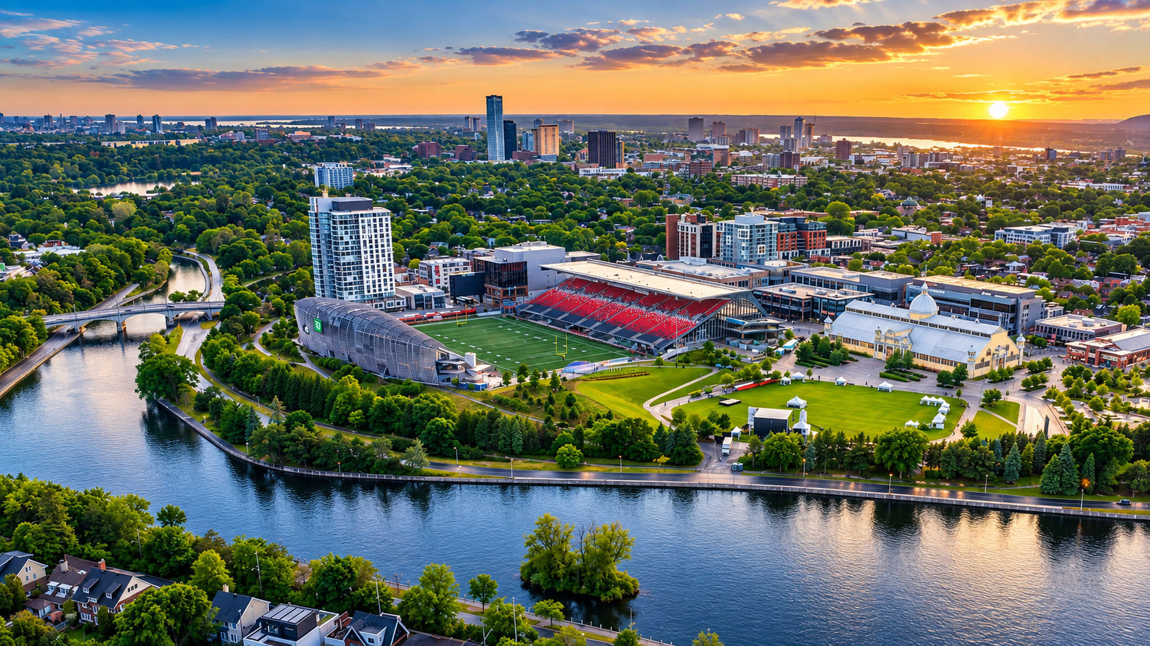 Lansdowne Park near The Glebe in Ottawa. Aerial view of a city with a river in the foreground, sports stadium, green parks, and residential and commercial buildings at sunset.