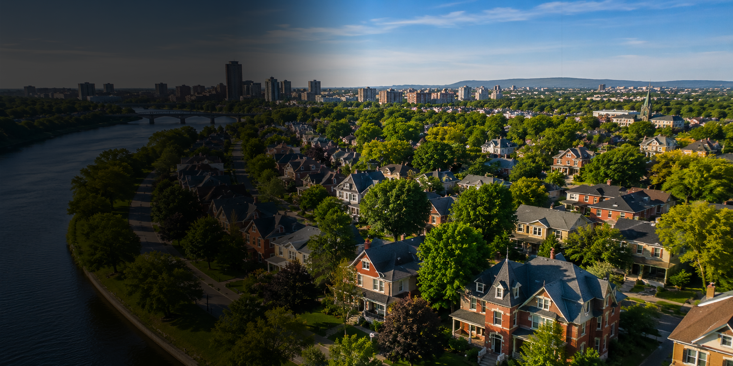 Aerial view of a city with a river on the left, green trees, houses, and high-rise buildings in the background under a partly cloudy sky.