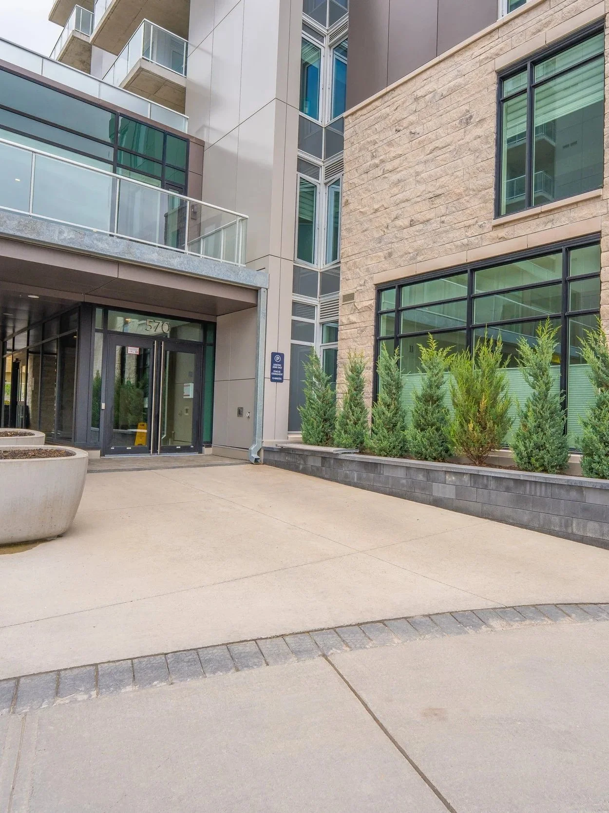 Modern apartment building entrance with glass doors, balcony railings, and large windows. Small evergreen plants line the building's exterior, and there is a large concrete planter in the foreground. The image shows a clean, urban residential area with a neutral color palette.