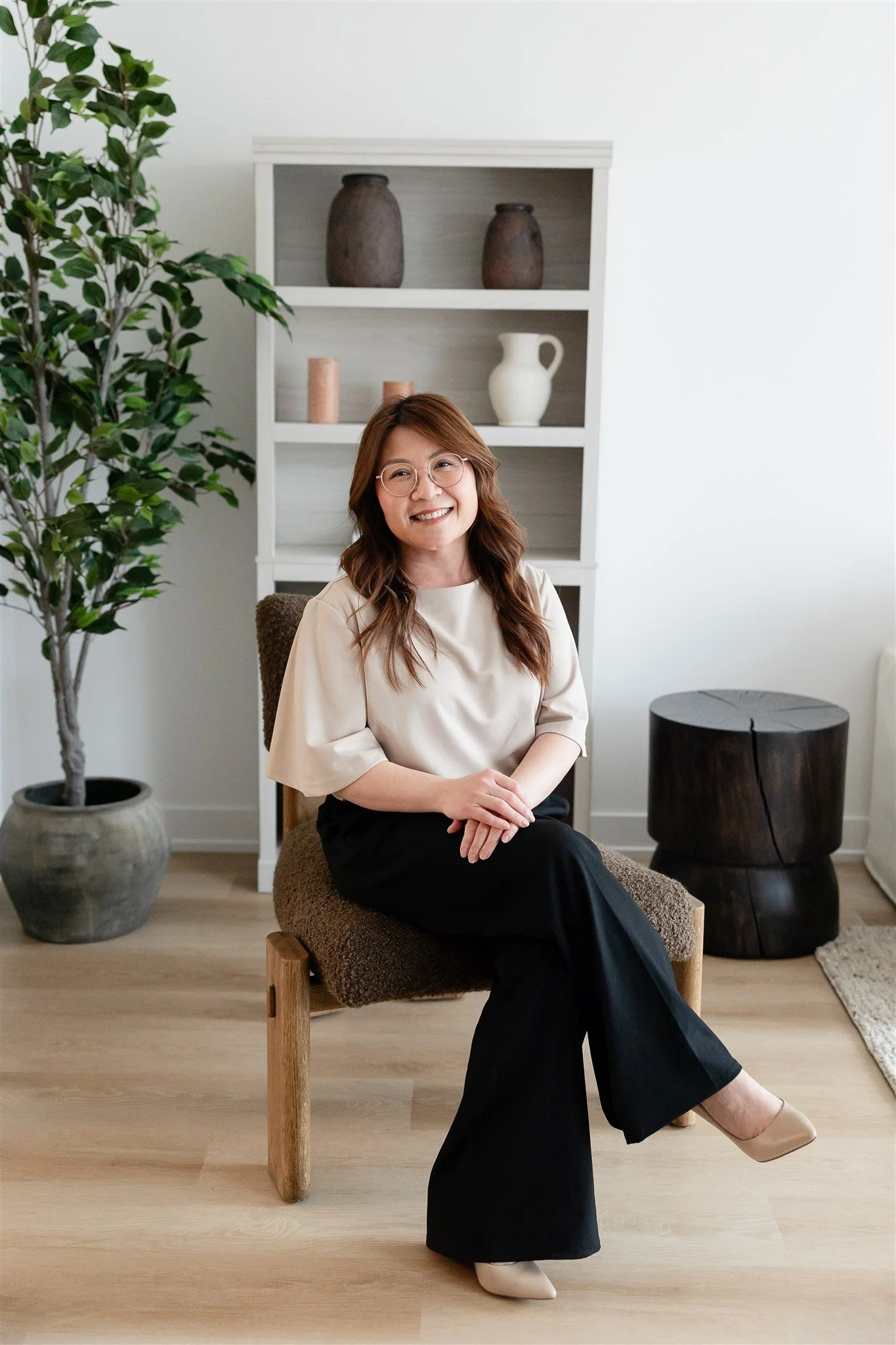A woman with long hair and glasses smiling, wearing a light-colored top, standing indoors in front of a dark-colored mantel with a round mirror on the wall.