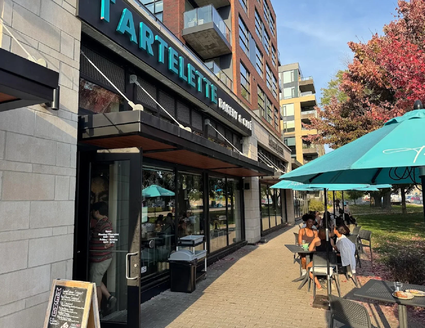 People sitting at outdoor tables with umbrellas in front of a restaurant called 'Artieletté Bakery & Coffee' on a city street with modern buildings and colorful trees.
