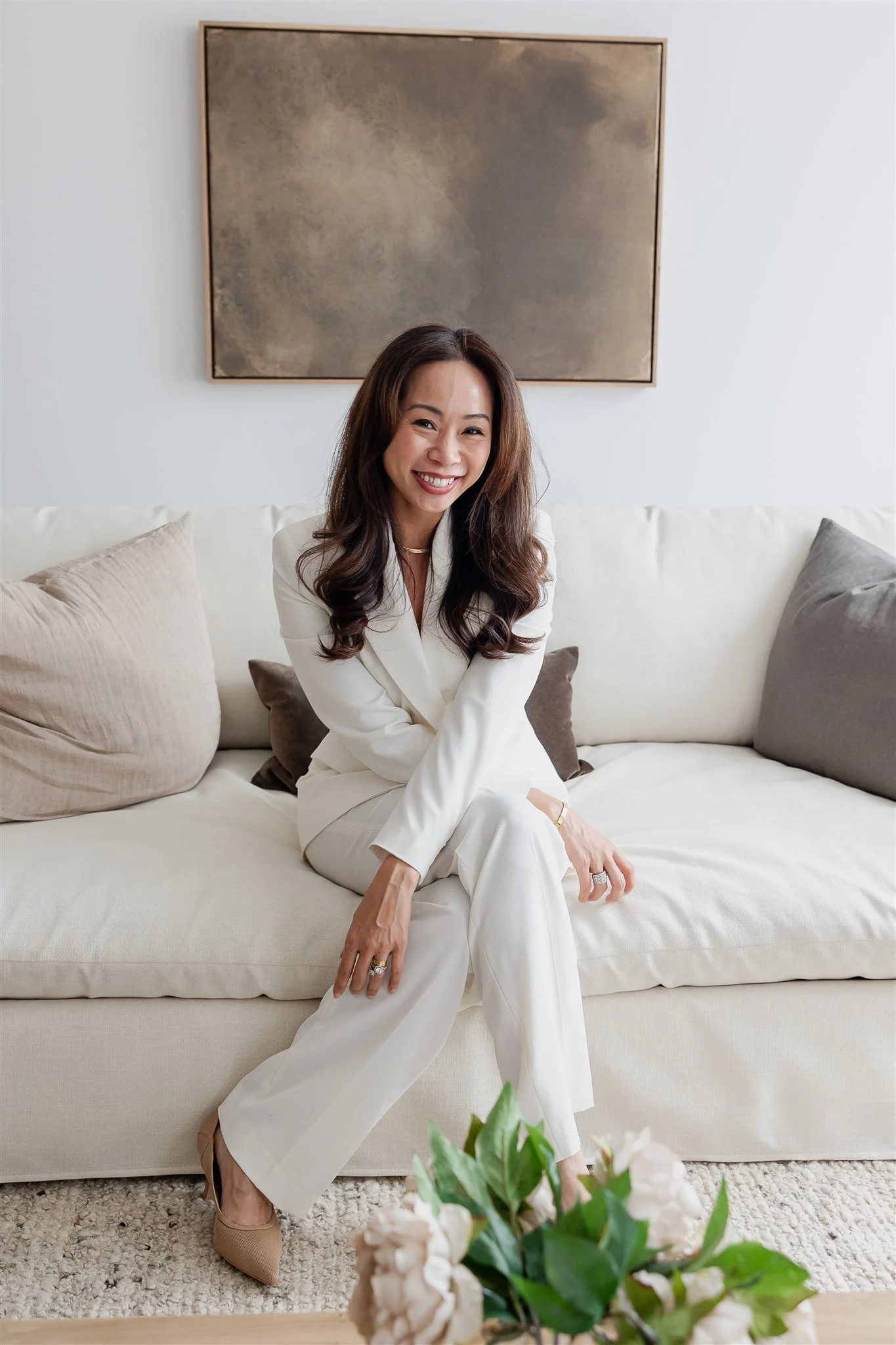 Smiling woman sitting on a chair, wearing white outfit, with a mirror in the background.