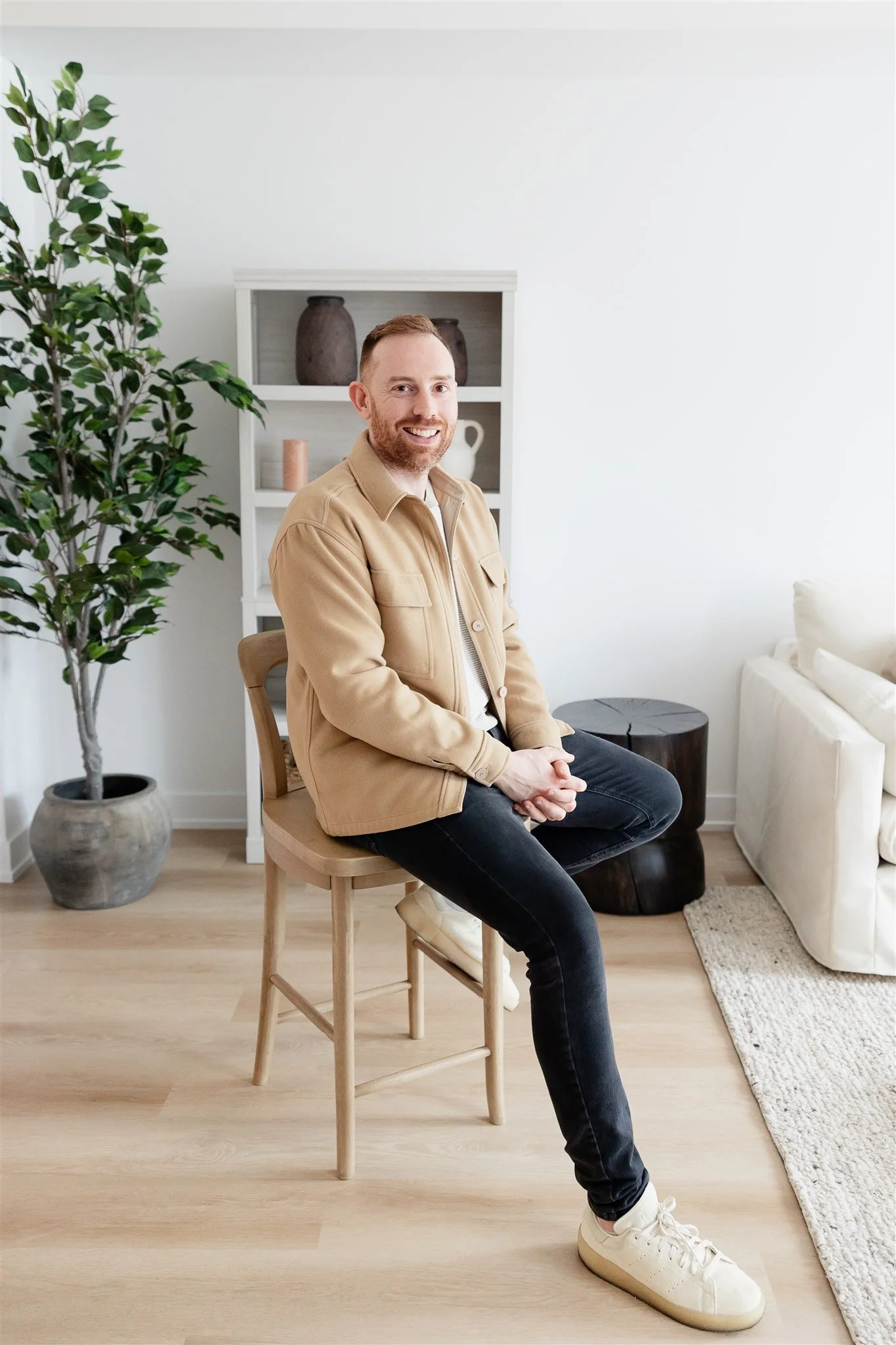 Man with a beard sitting indoors, wearing a tan jacket, with a staircase in the background.