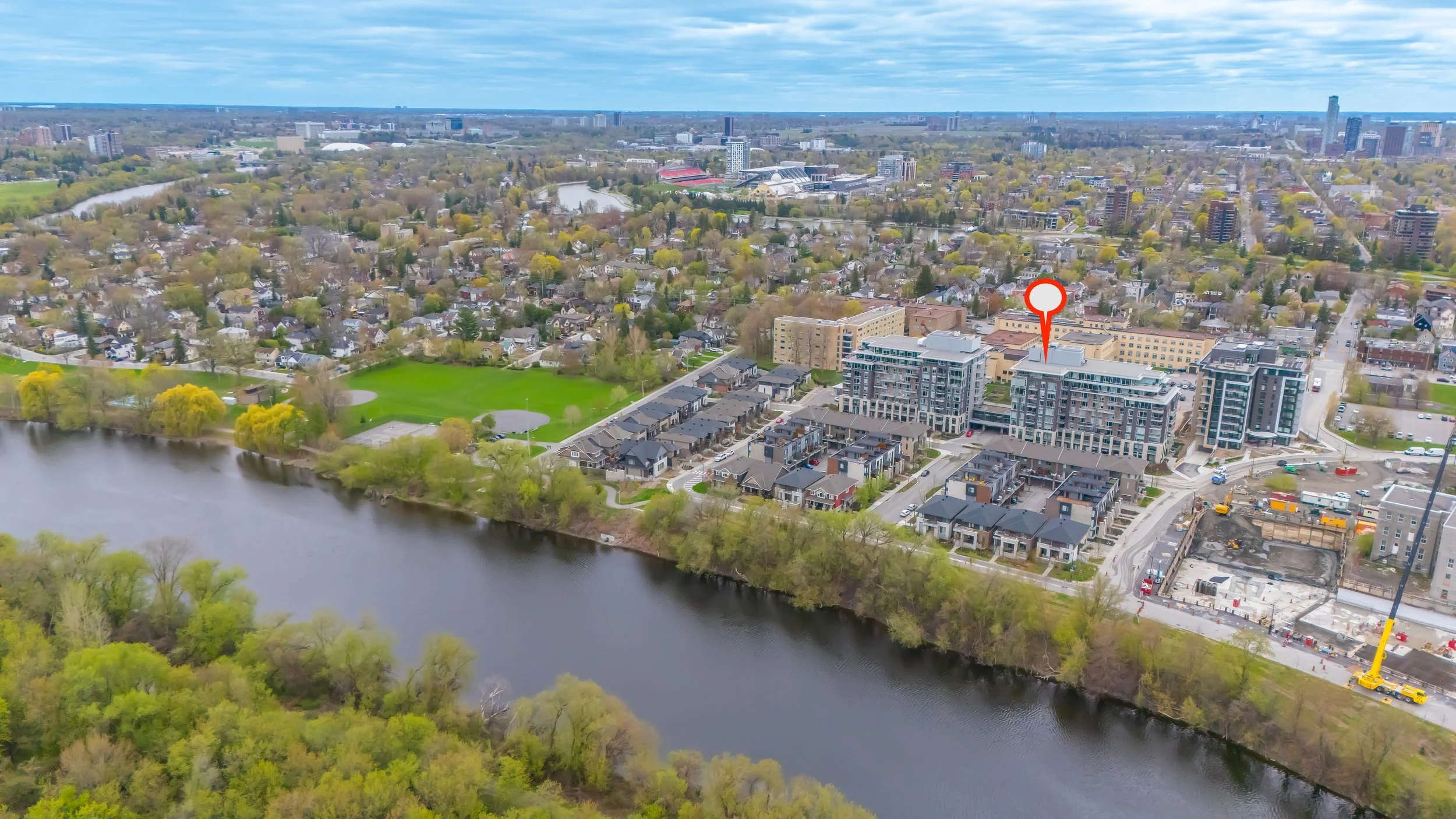 An aerial view of a cityscape featuring a river with green trees along its banks in the foreground, residential houses, apartment buildings, and a green park area, with a skyline of high-rise buildings, a stadium, and a cloudy sky in the background.