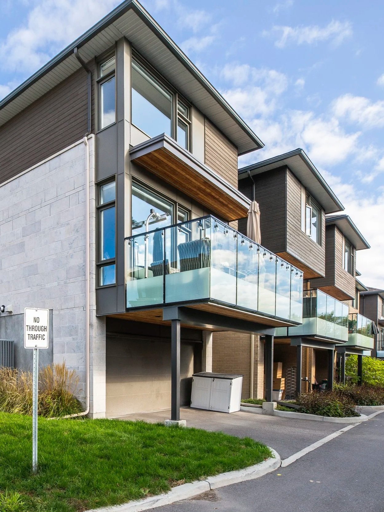 Modern multi-story apartment building with glass balconies, stucco and wood siding, situated in a residential area with a small lawn, a 'No Through Traffic' sign, and a clear blue sky.