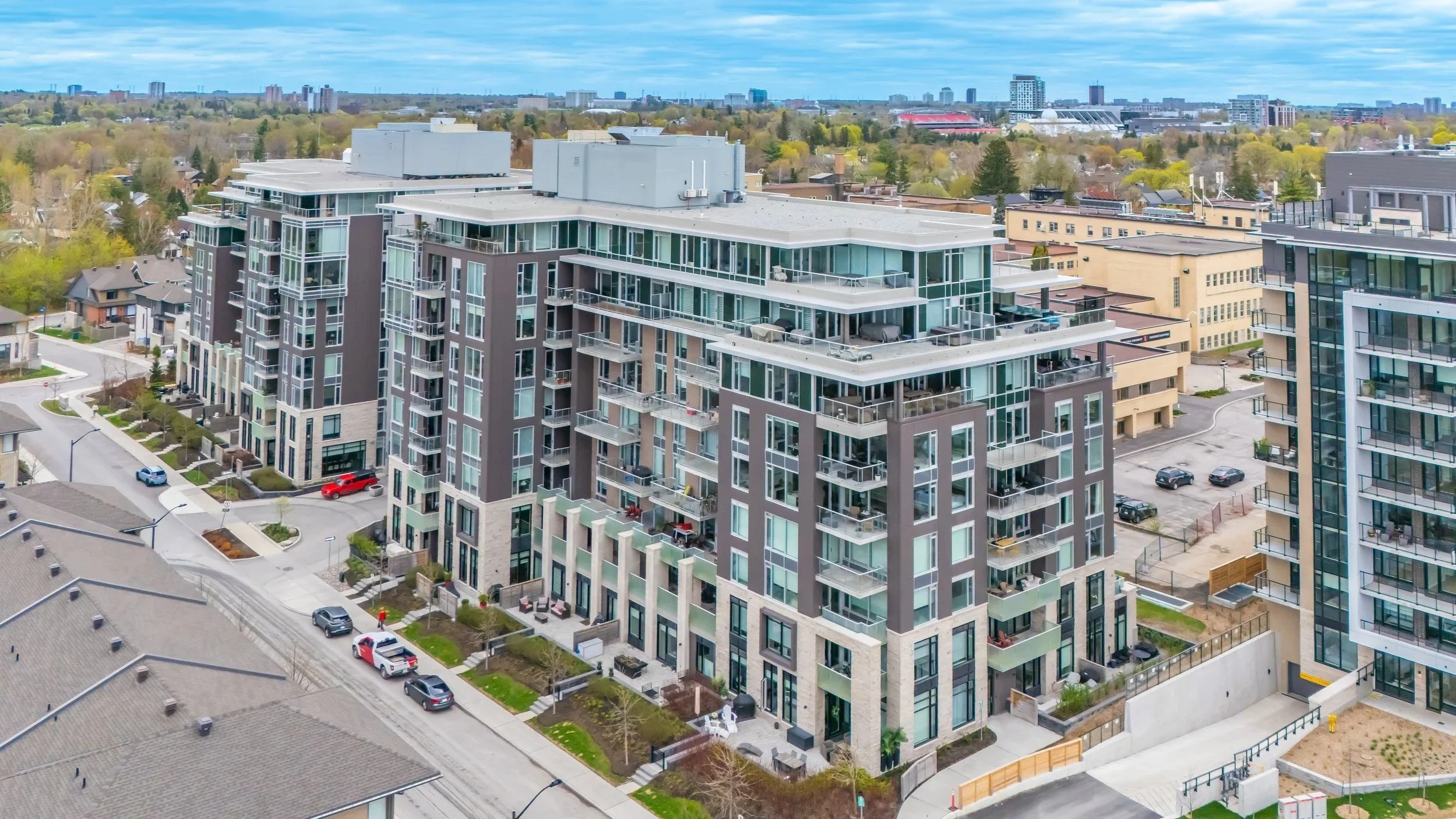 Aerial view of modern apartment buildings with glass balconies in an urban area within Greystone Village Ottawa, with streets, parked cars, and green trees.