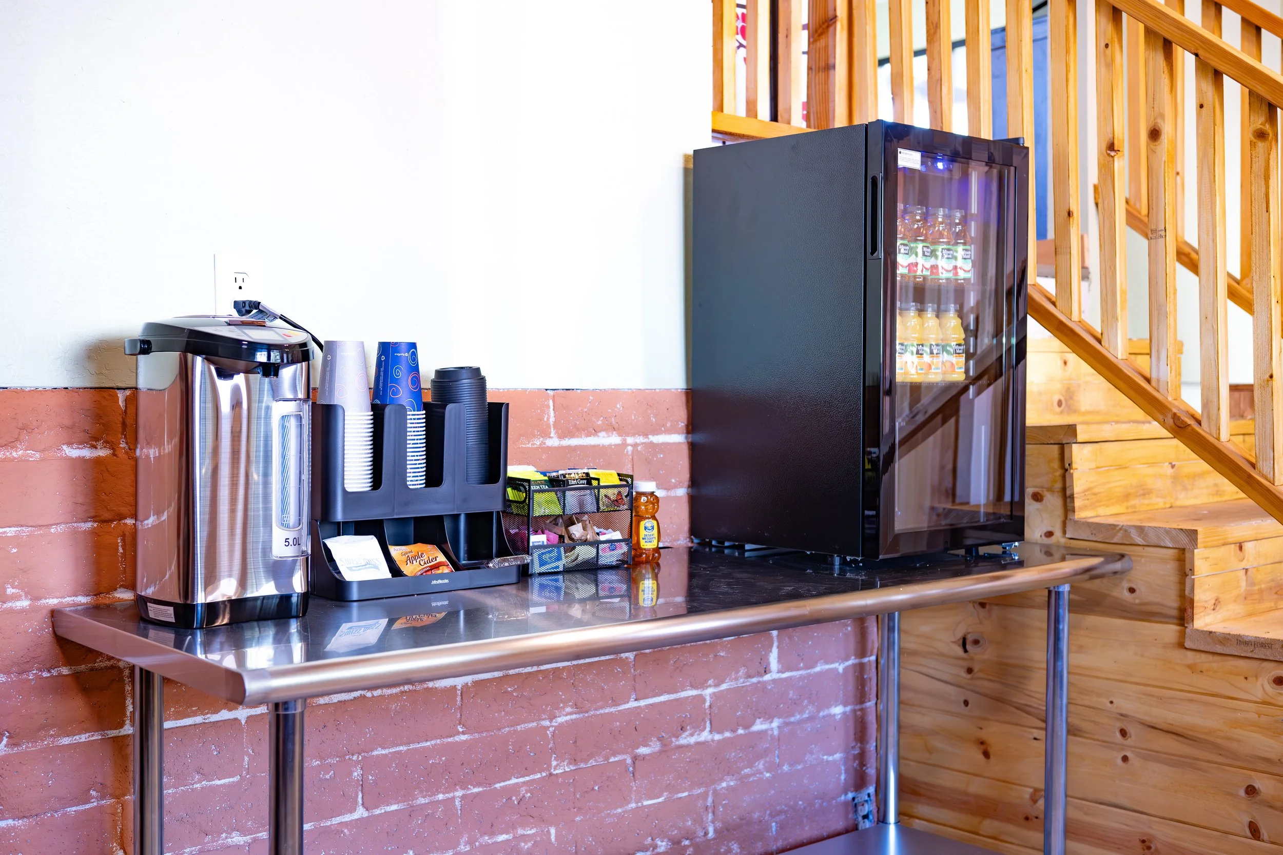 Coffee station with a stainless steel hot water boiler, disposable cups, coffee pods, tea packets, and a small fridge on a wooden counter next to brick and wood walls.