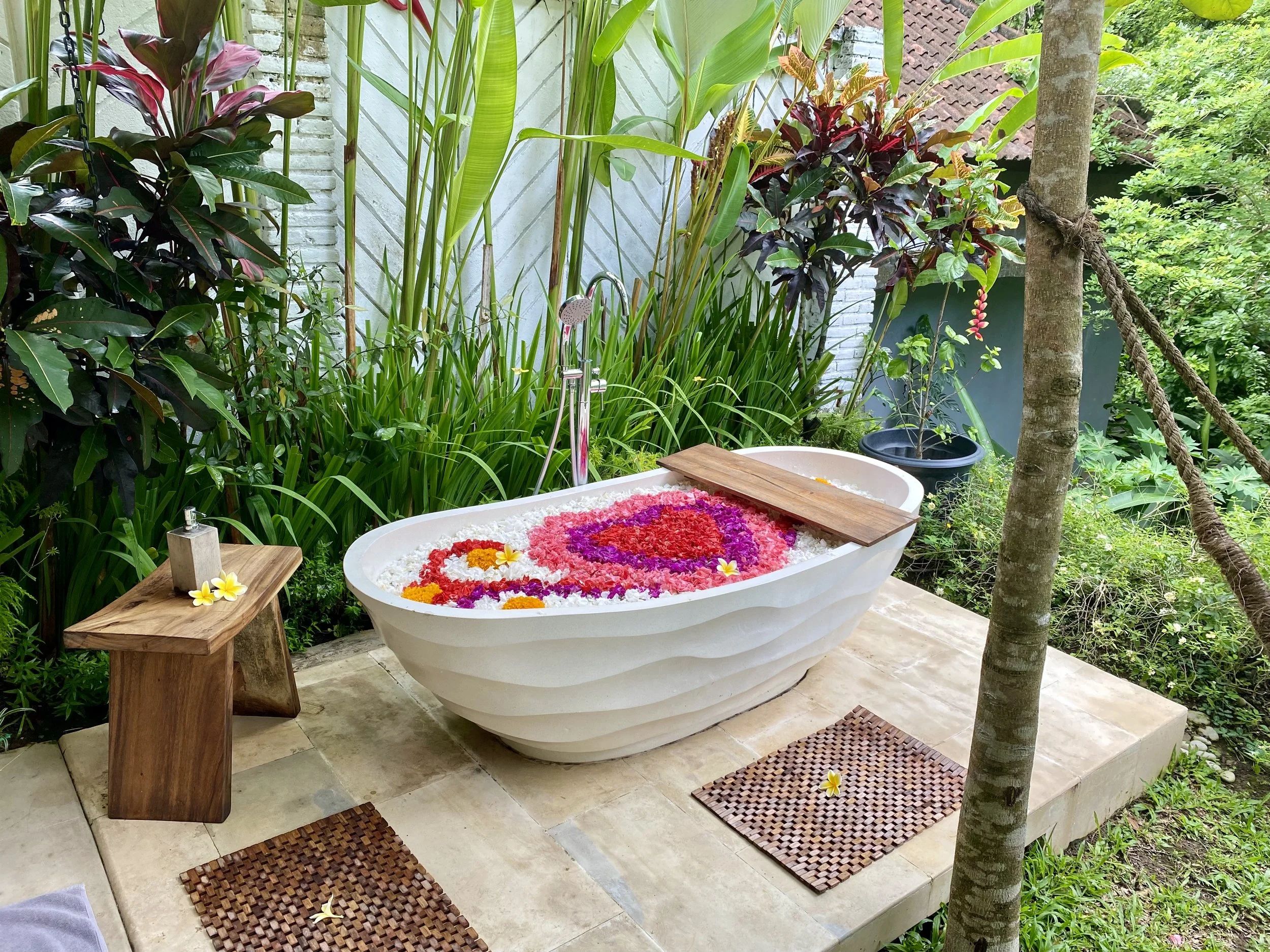 Outdoor garden with a white bathtub filled with flower petals and floating yellow flowers, wooden covers, and green lush plants and trees around.