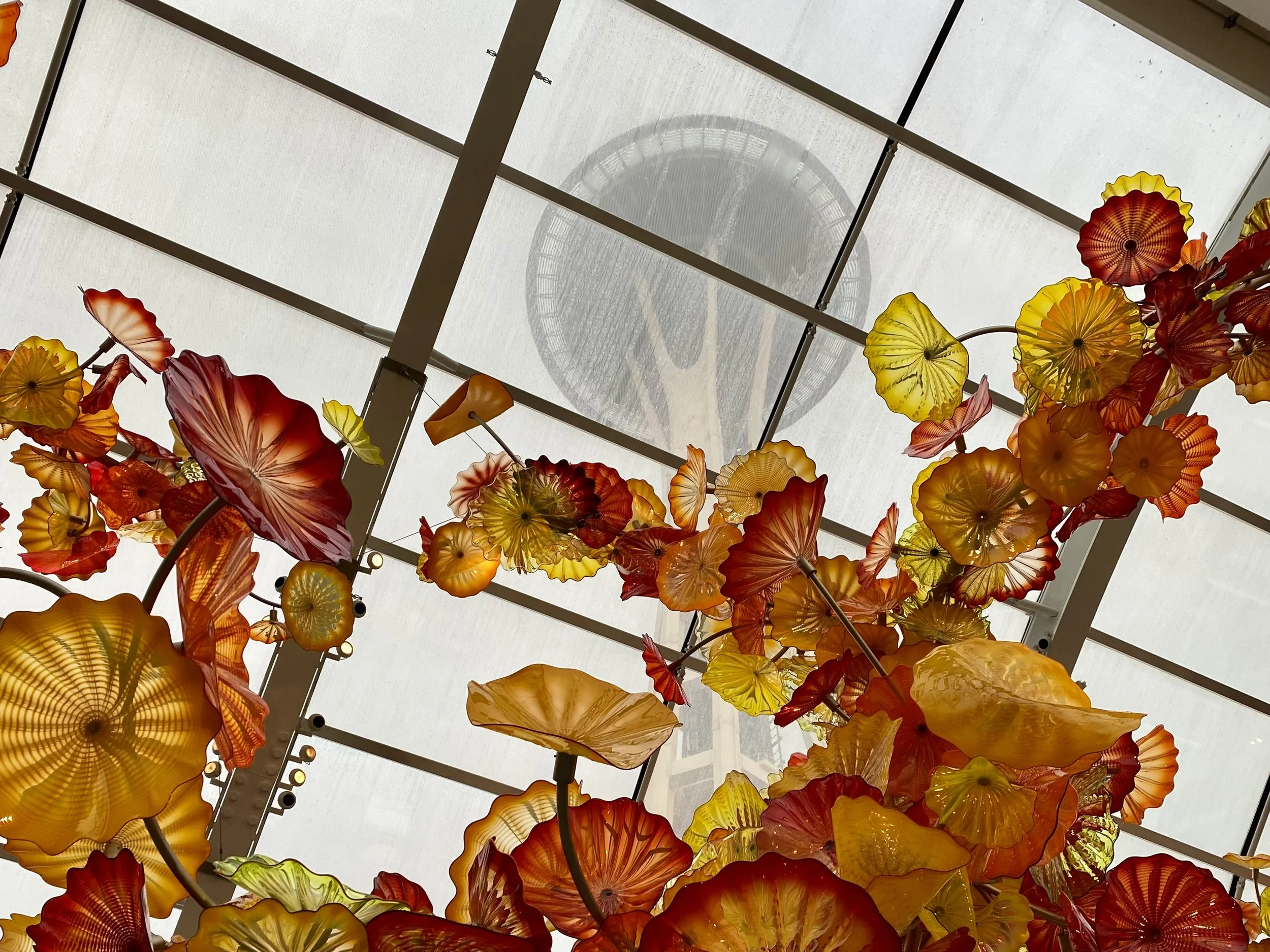 Colorful glass art installation hanging from the ceiling with yellow, orange, and red flower-like shapes, viewed from below.