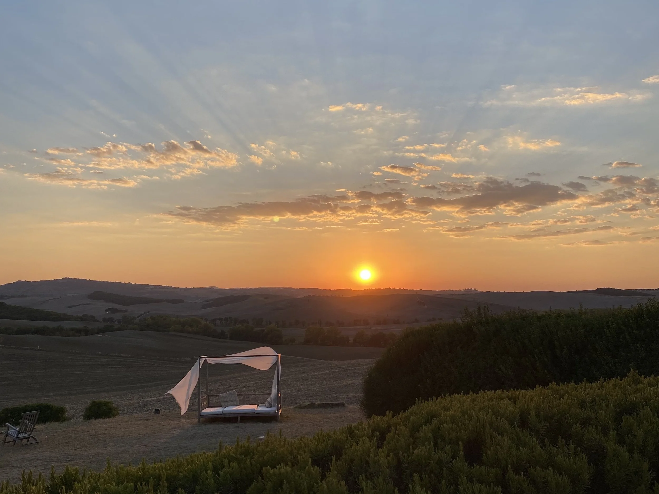 A scenic landscape of rolling hills during sunset with a partly cloudy sky. In the foreground, there is a cozy outdoor bed with canopy, positioned on a grassy area along with a bench to the left. The sky is illuminated with warm colors, and the sun is near the horizon.