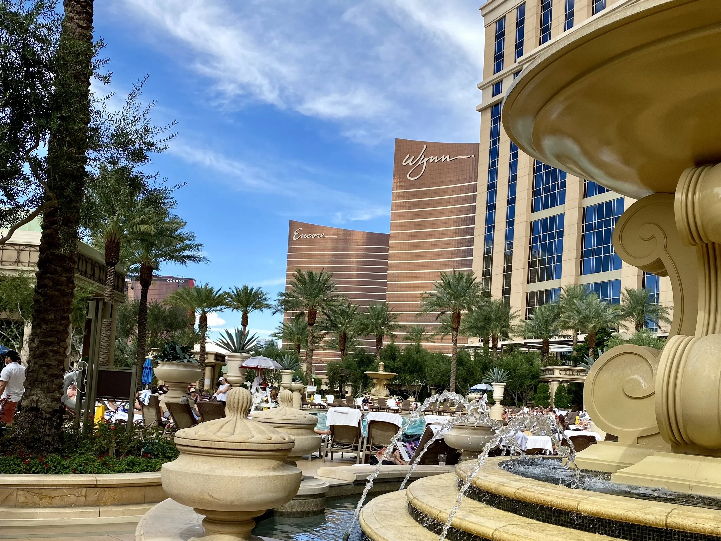View of a Las Vegas hotel pool area with palm trees, lounge chairs, umbrellas, and fountains, with the Wynn and Encore hotel towers in the background under a partly cloudy sky.
