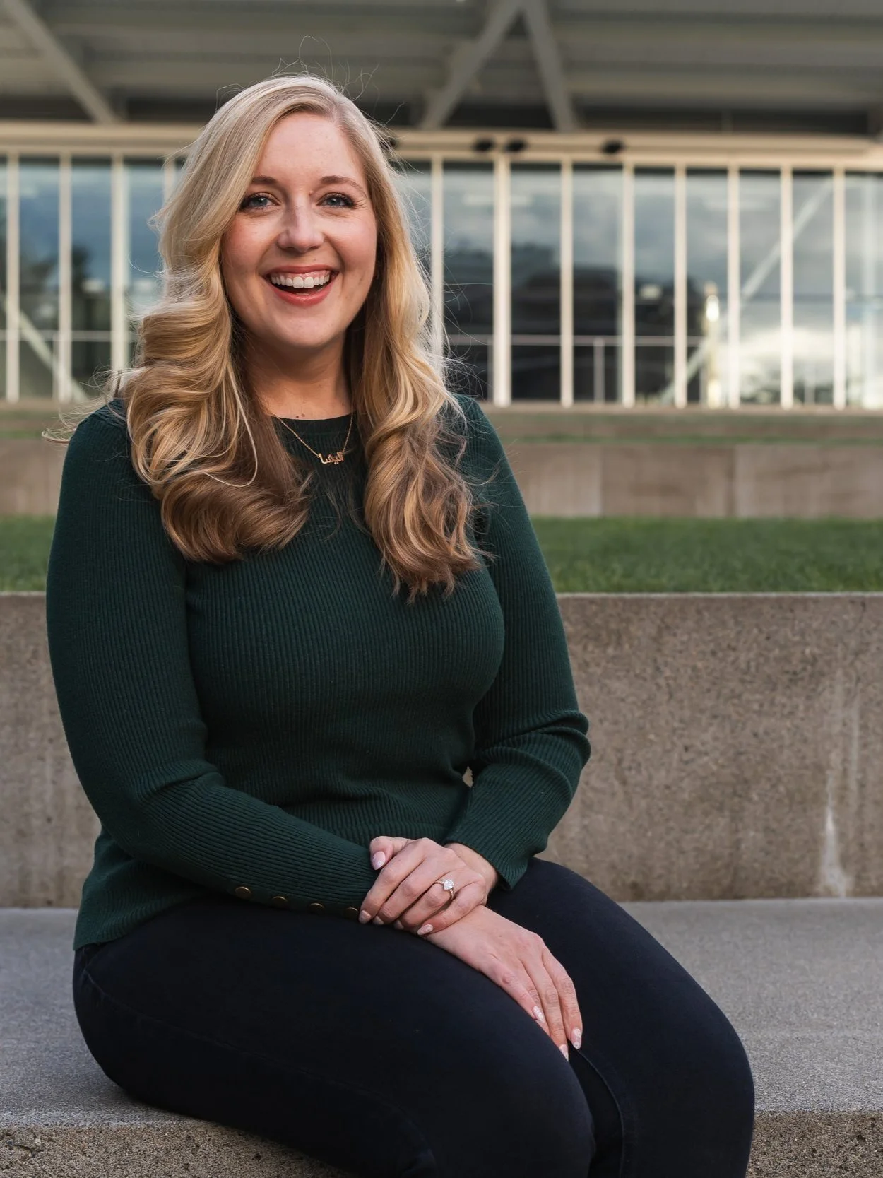 A smiling woman with long blonde hair sitting outdoors on a concrete ledge in front of a modern glass building.
