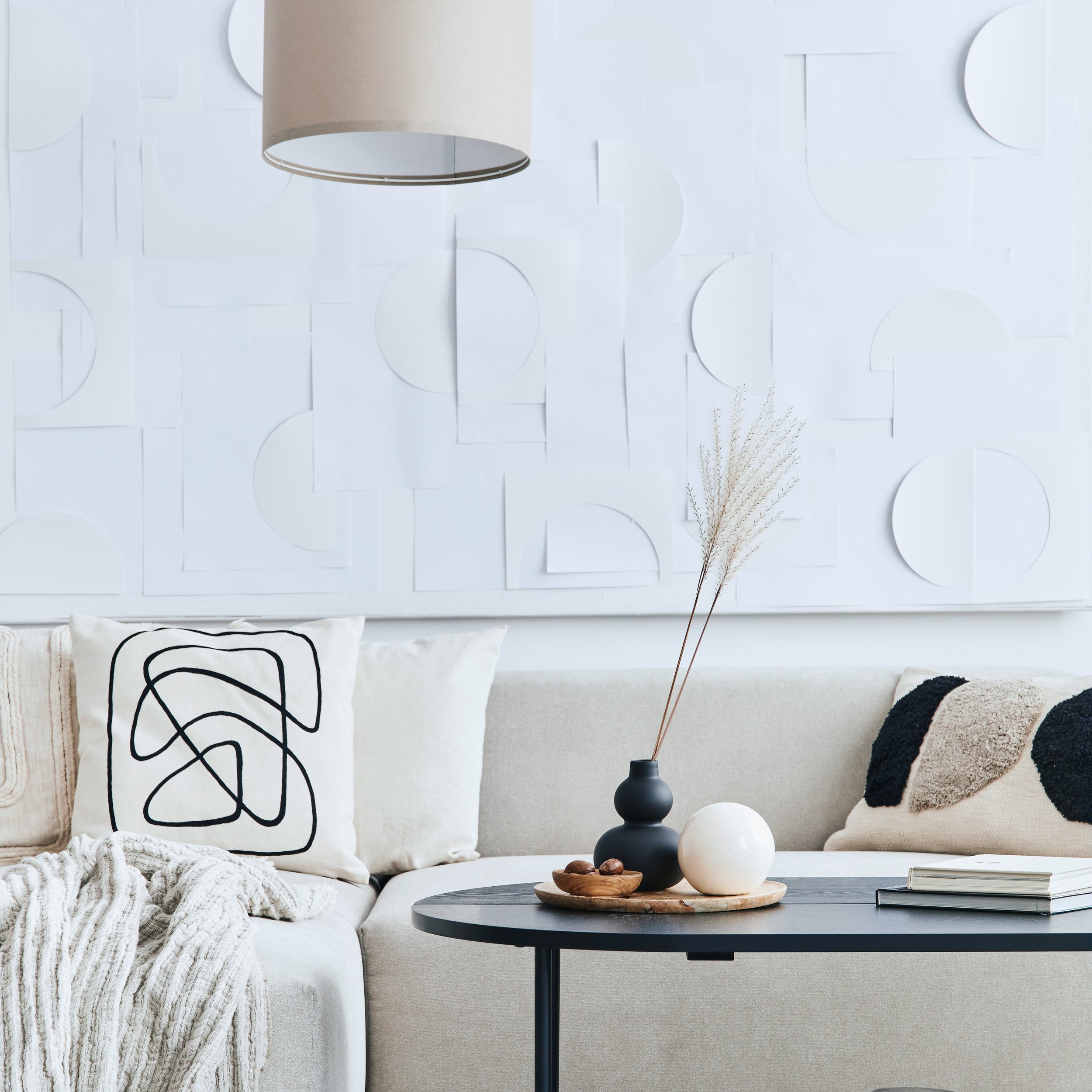 A modern living room with white textured wall art, a black round side table, a white couch with decorative pillows, a beige blanket, and a black vase with dried pampas grass.