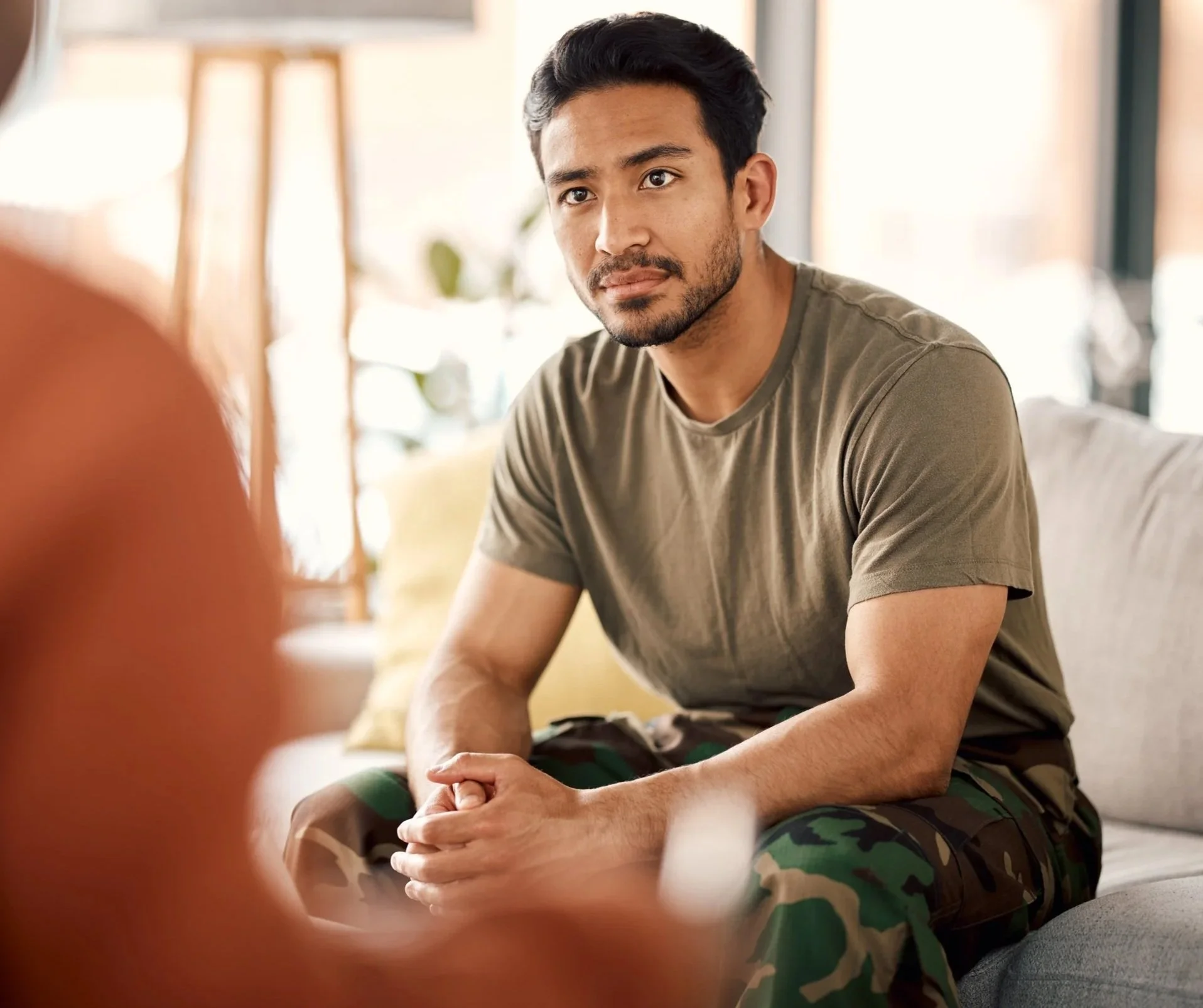A man sitting with a serious expression, wearing a gray T-shirt and camouflage pants, in a bright, modern room with a blurred background.