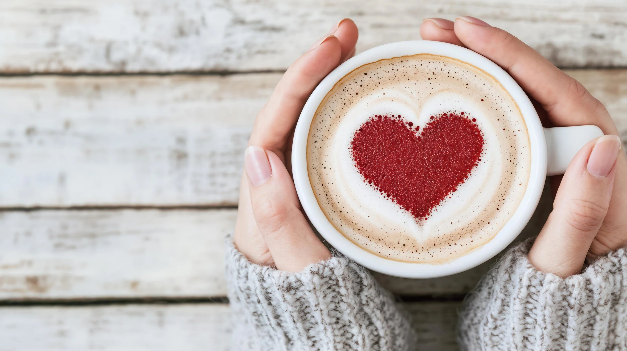 Person holding a white mug filled with frothy coffee decorated with a red heart design.