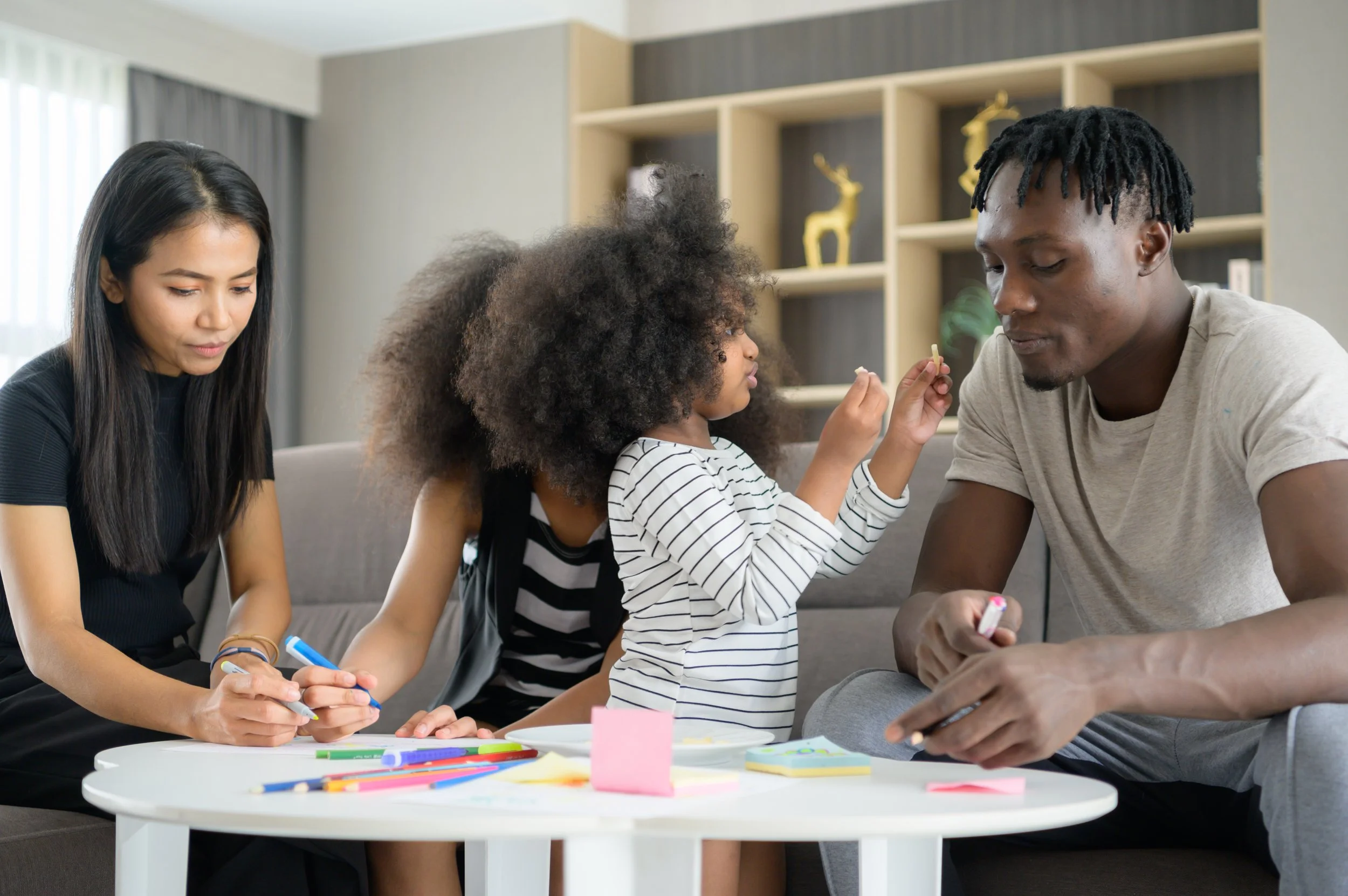 A group of four people, including two women and two children, sit around a white table engaging in an activity with colorful markers and paper in a living room with bookshelves and decorative items in the background.