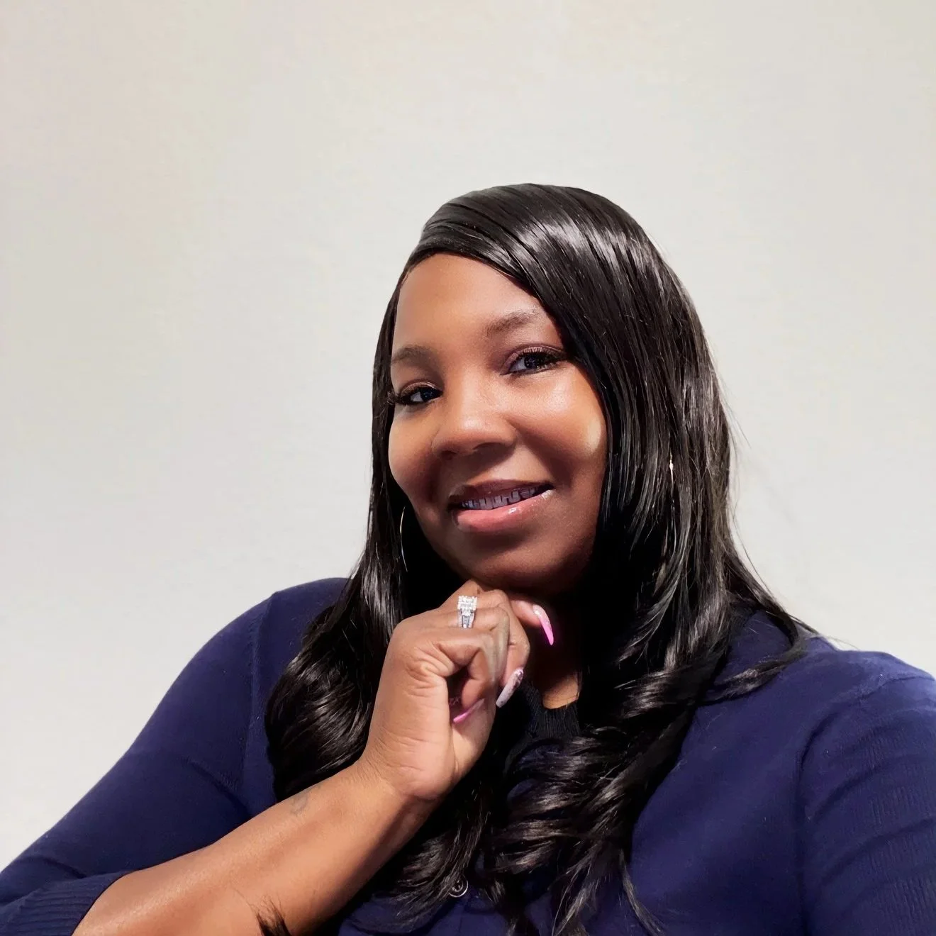 A woman with long black hair and hoop earrings, smiling and resting her chin on her hand, posing against a plain white wall.