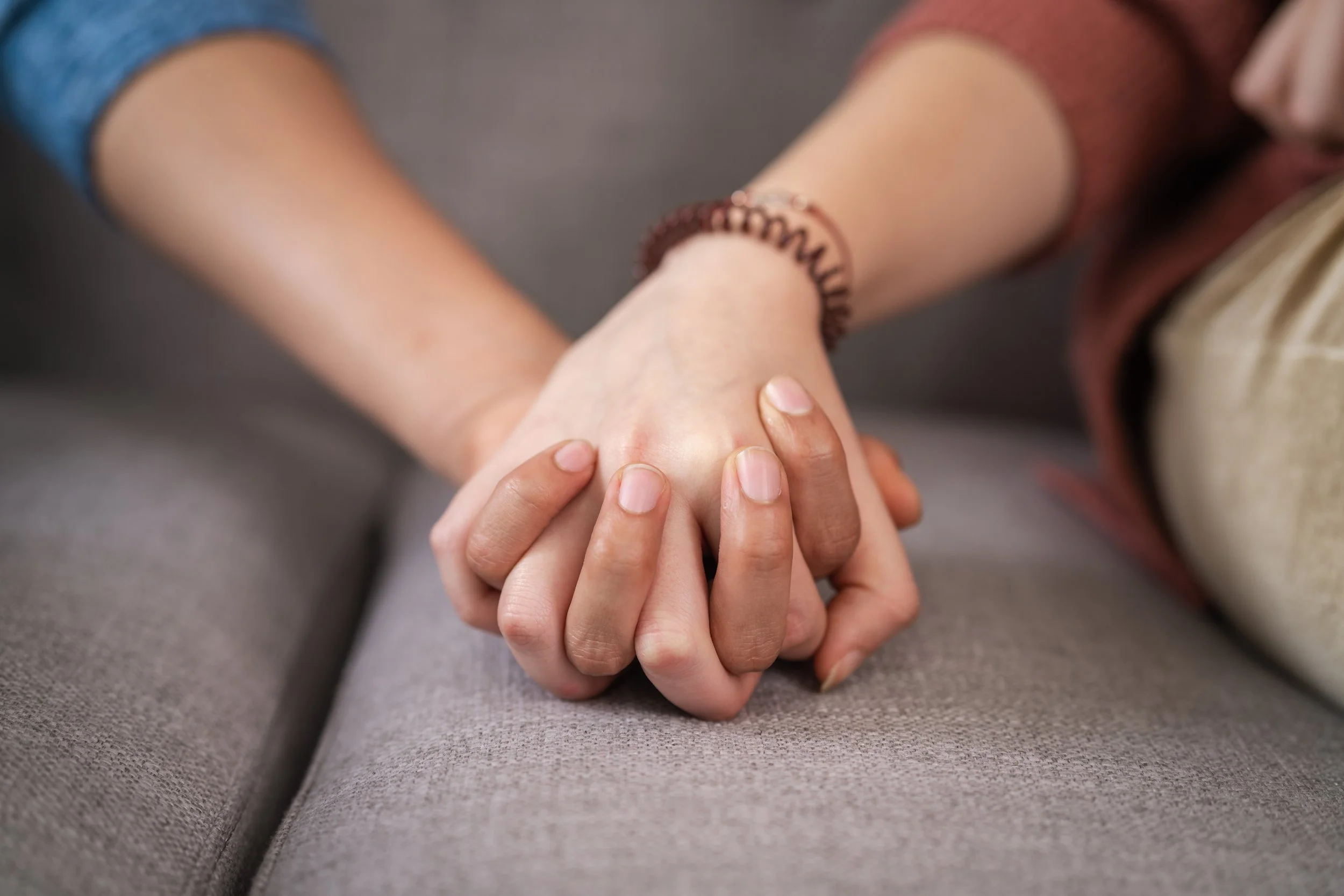 Close-up of two people holding hands, one with a beaded bracelet, on a gray fabric surface.