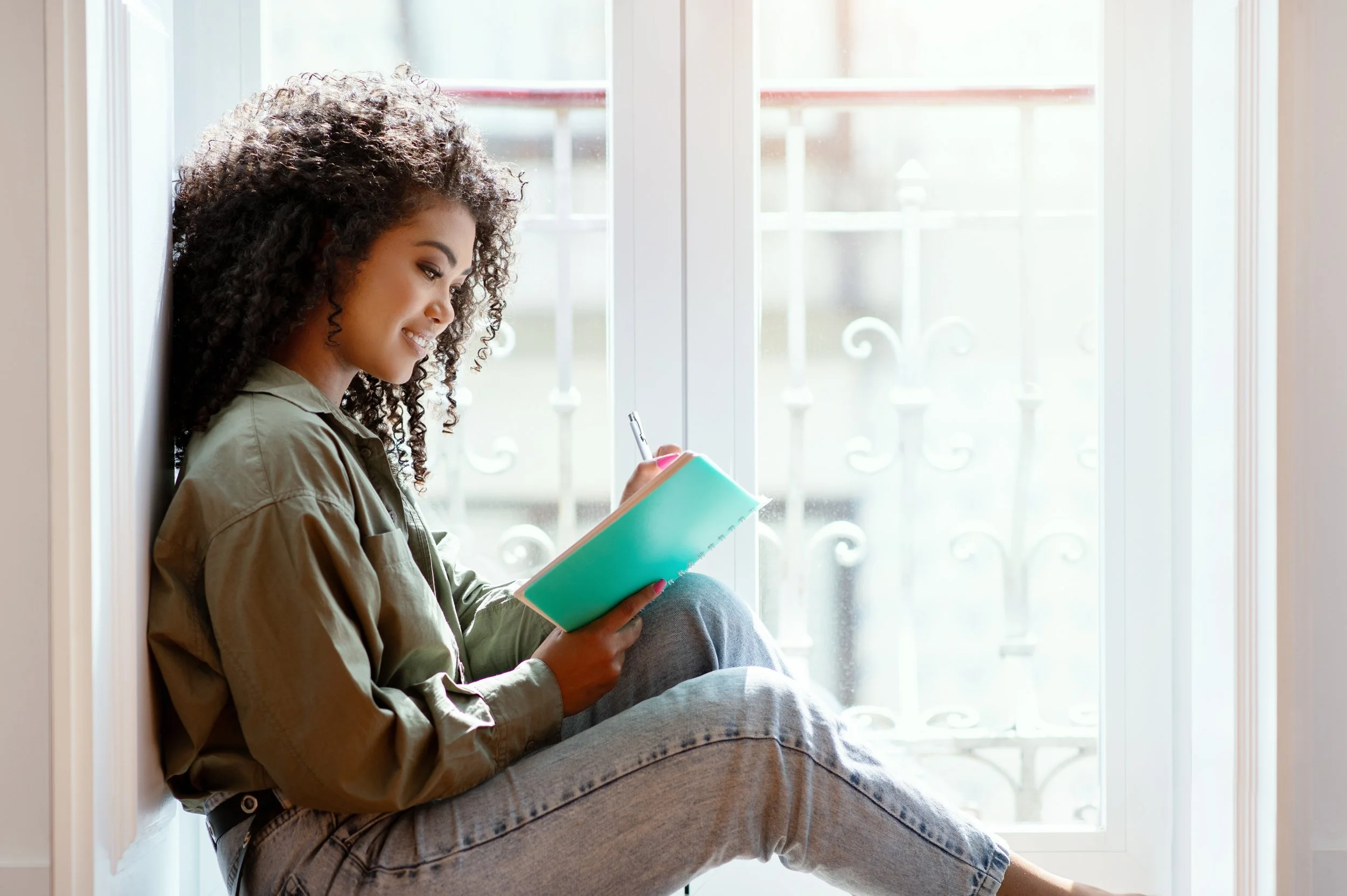 Young woman with curly hair sitting by a large window, smiling while writing in a notebook.