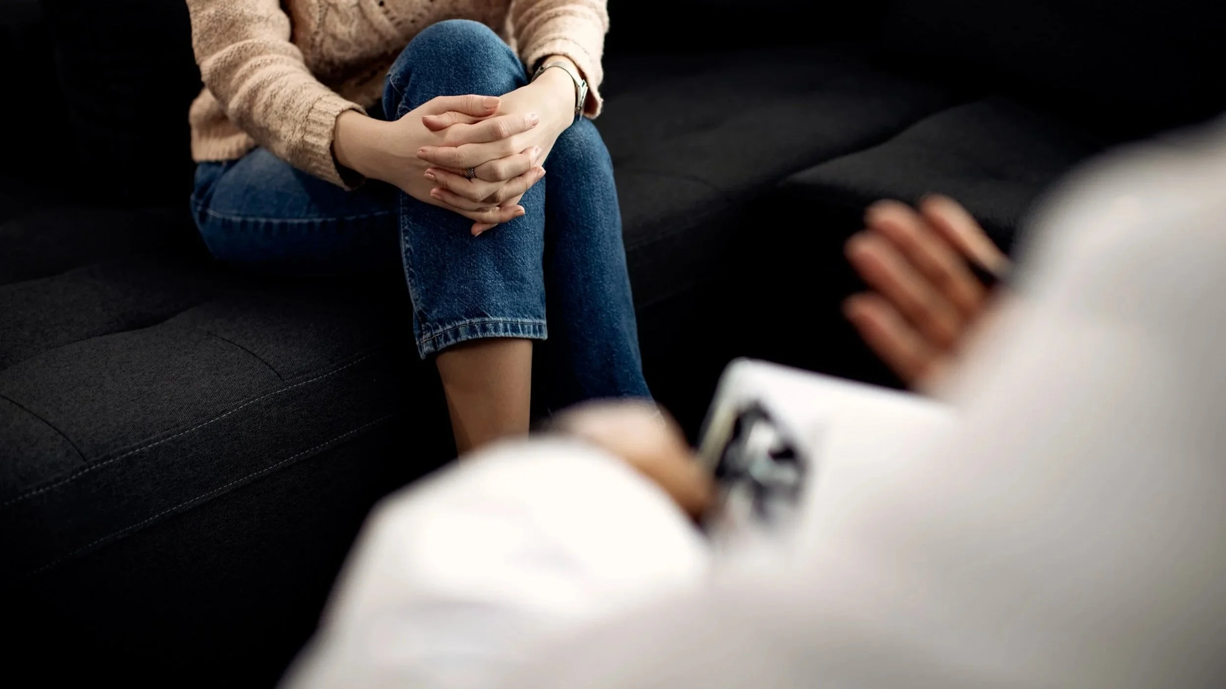 Person wearing a beige sweater and blue jeans sitting on a black sofa, with hands clasped together, during a conversation or therapy session.