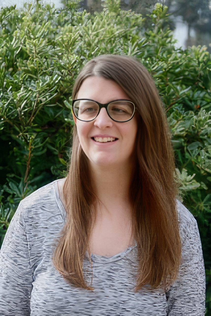 A young woman with long brown hair wearing glasses and a gray patterned shirt, smiling outdoors in front of green shrubbery.