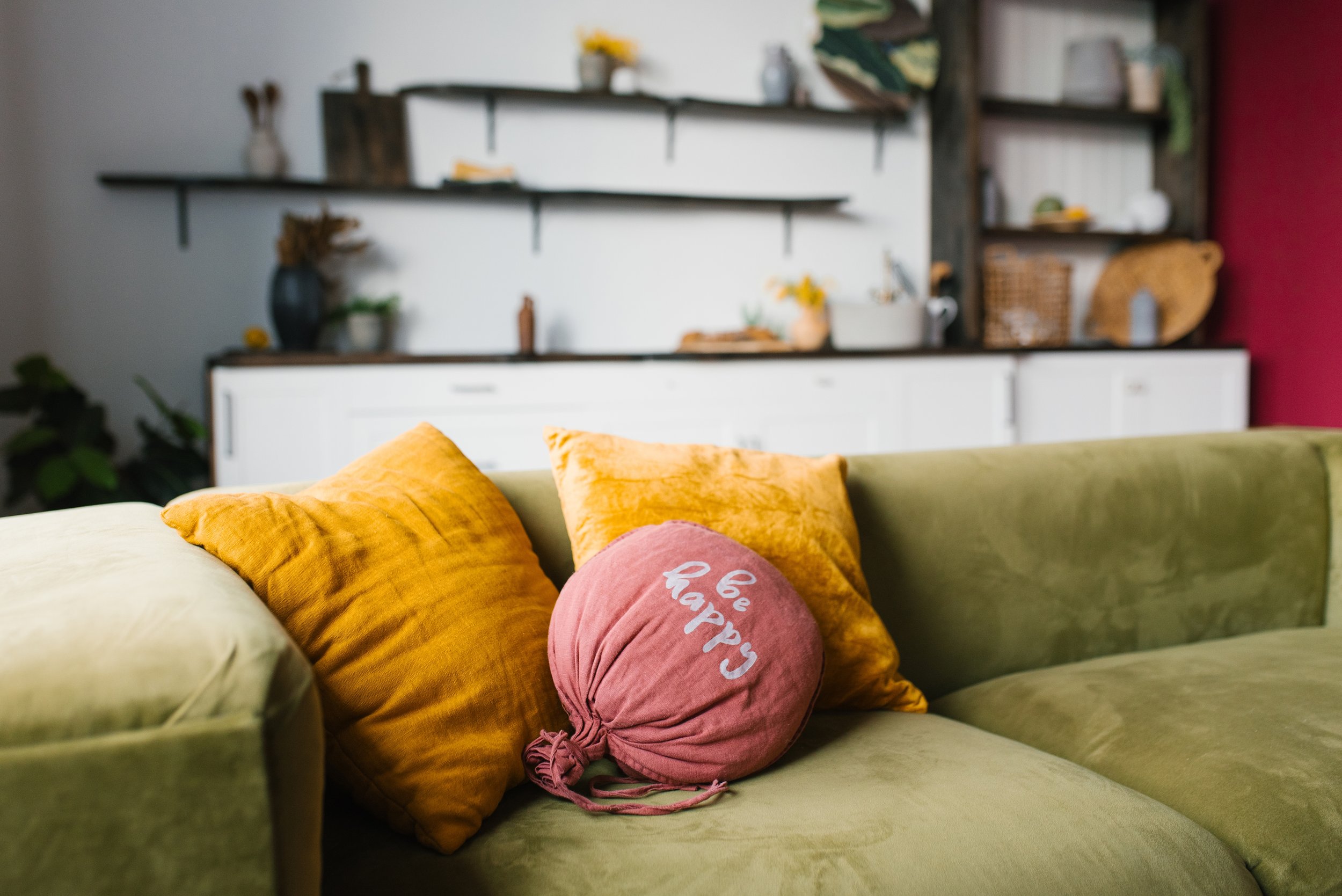A green sofa with mustard yellow and pink pillows, including a round pink pillow with the words 'be happy' written on it. In the background, a wall-mounted black shelving unit holds various decor items, including vases, plants, and baskets. The wall behind the shelves is white with part of a deep pink wall visible on the right side.