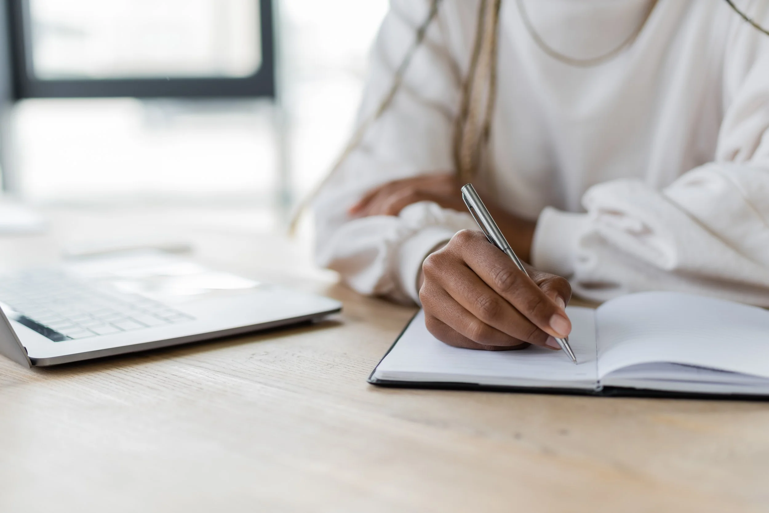 A person writing in a notebook with a pen, a laptop and a window in the background.