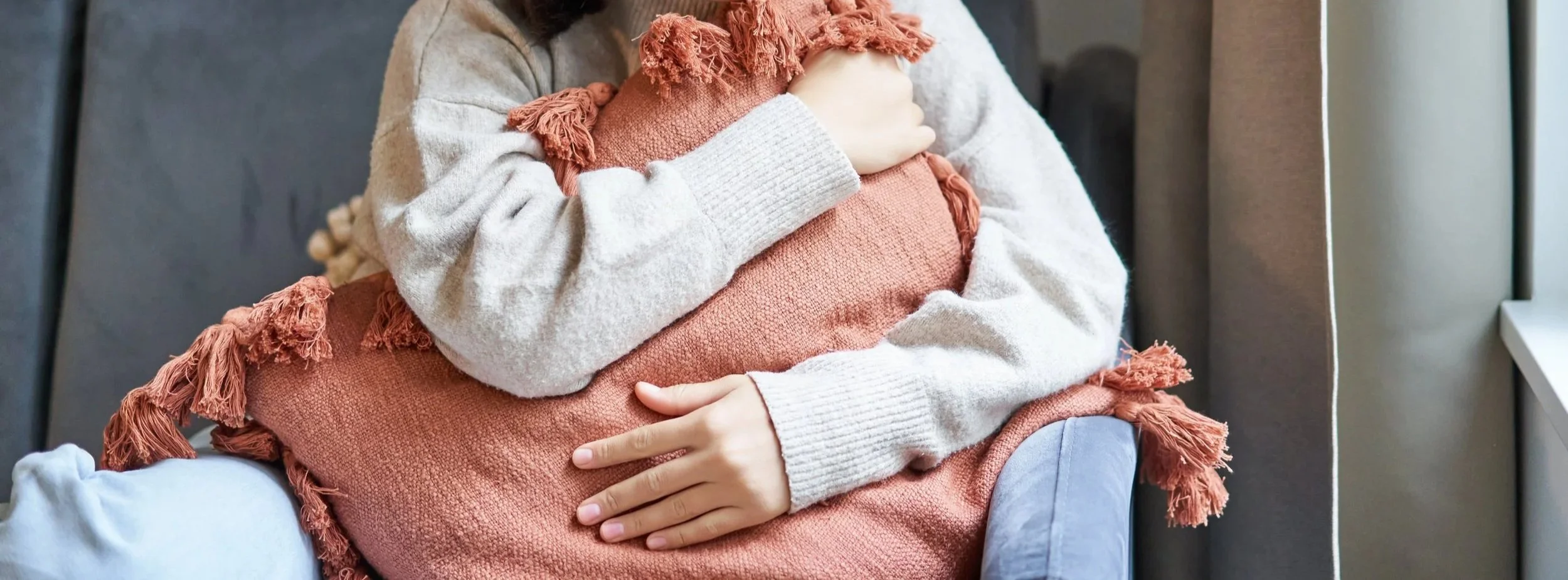 A person sitting on a gray chair, hugging a rust-colored throw pillow with tassels, while wearing a cozy beige sweater and light blue pants.